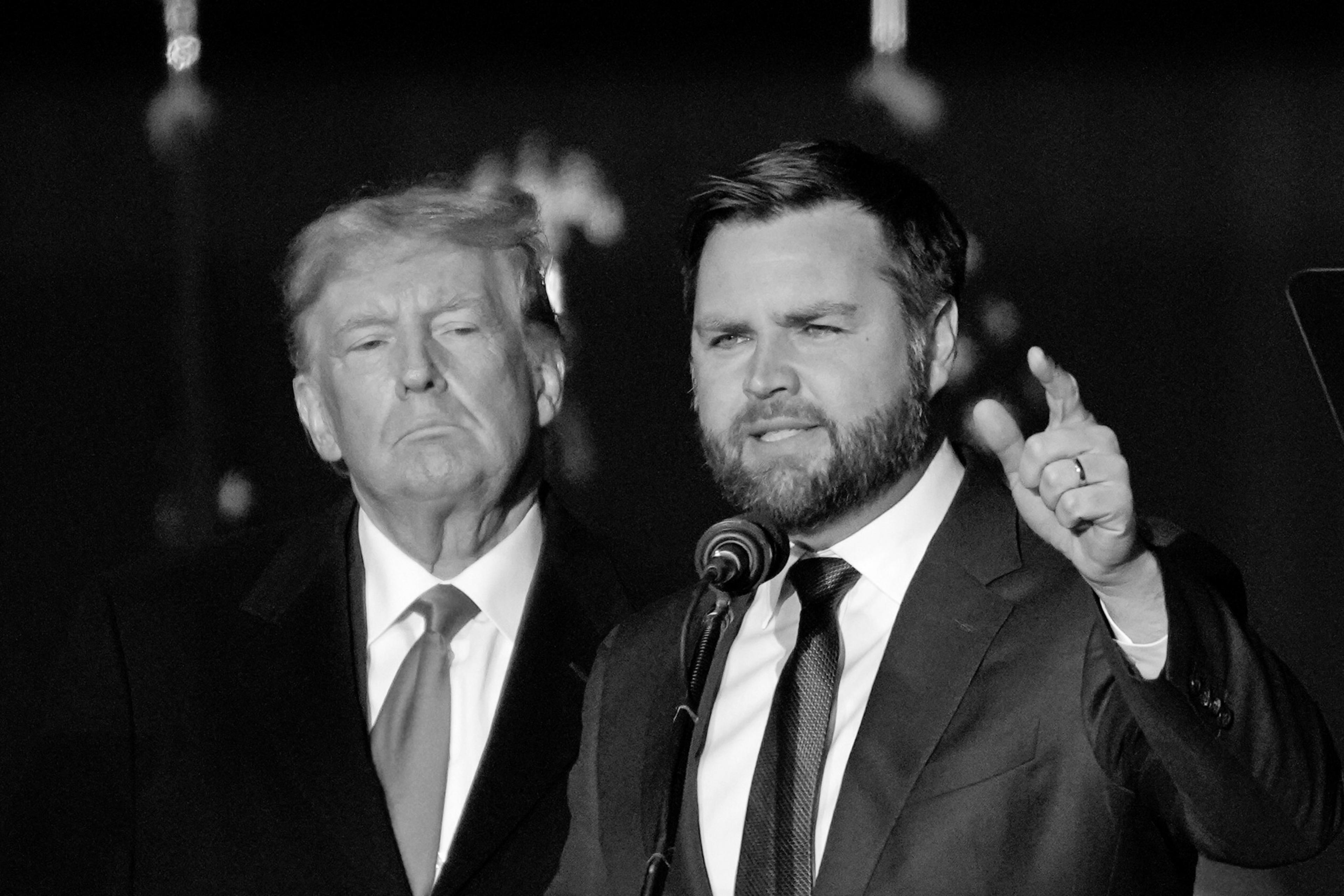 Two men in suits stand together. The man on the left looks forward with a serious expression, while the man on the right speaks into a microphone, gesturing with one hand.