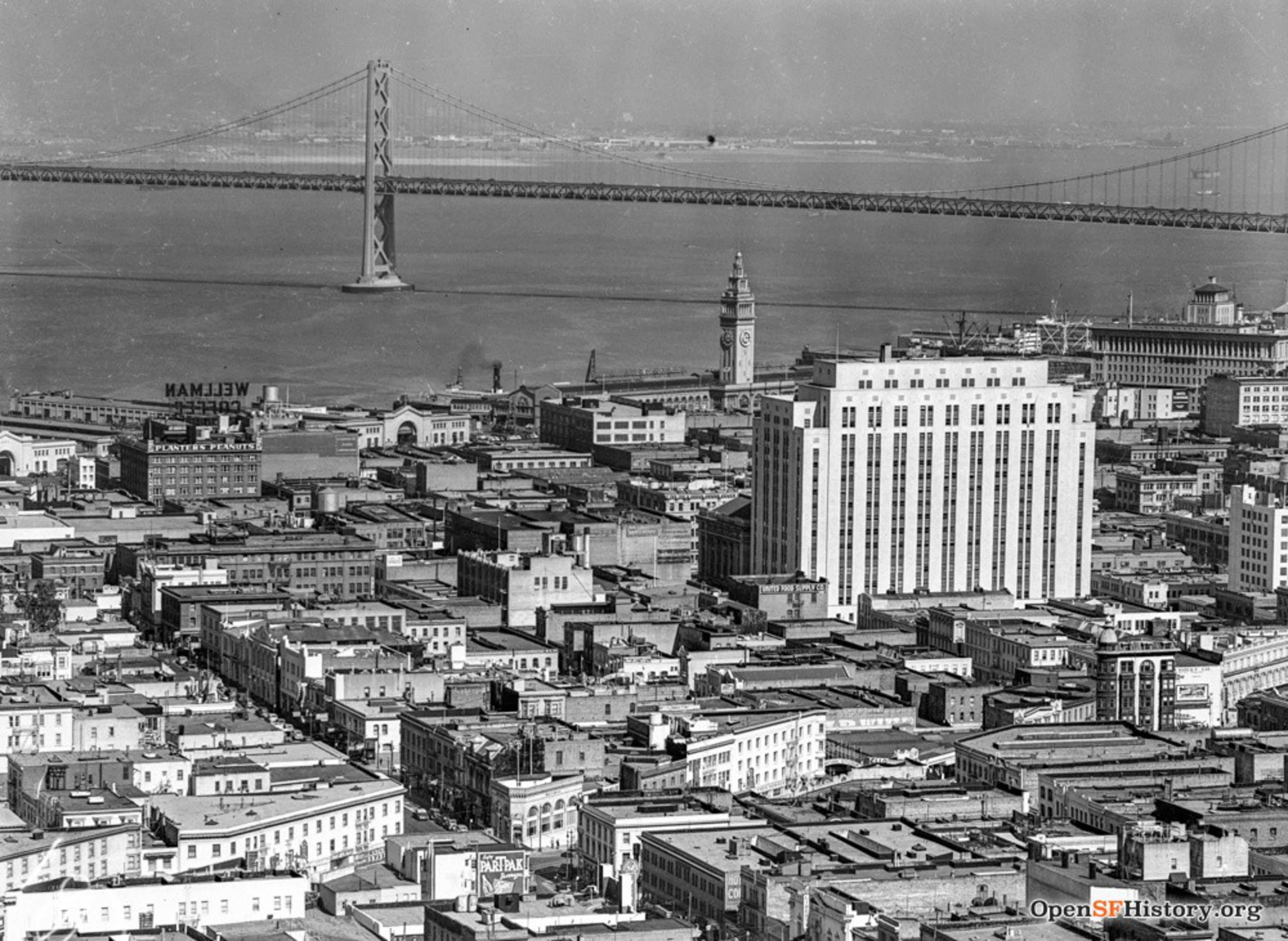 A black-and-white cityscape with numerous buildings, a tall clock tower near the waterfront, and a suspension bridge spanning across the water.