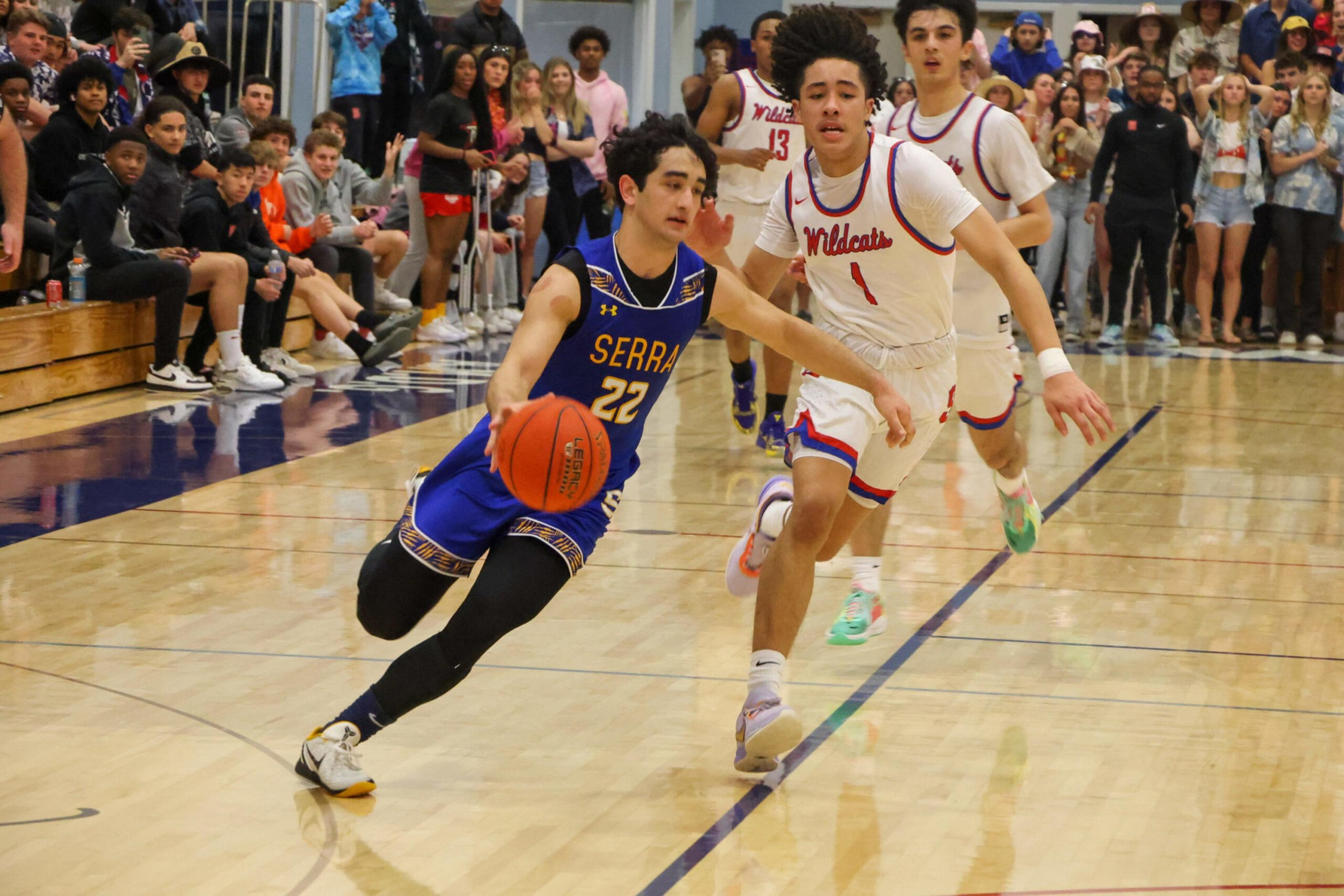 A basketball player in a blue "Serra" uniform dribbles past two defenders in white "Wildcats" jerseys on a gym court with a crowd watching.