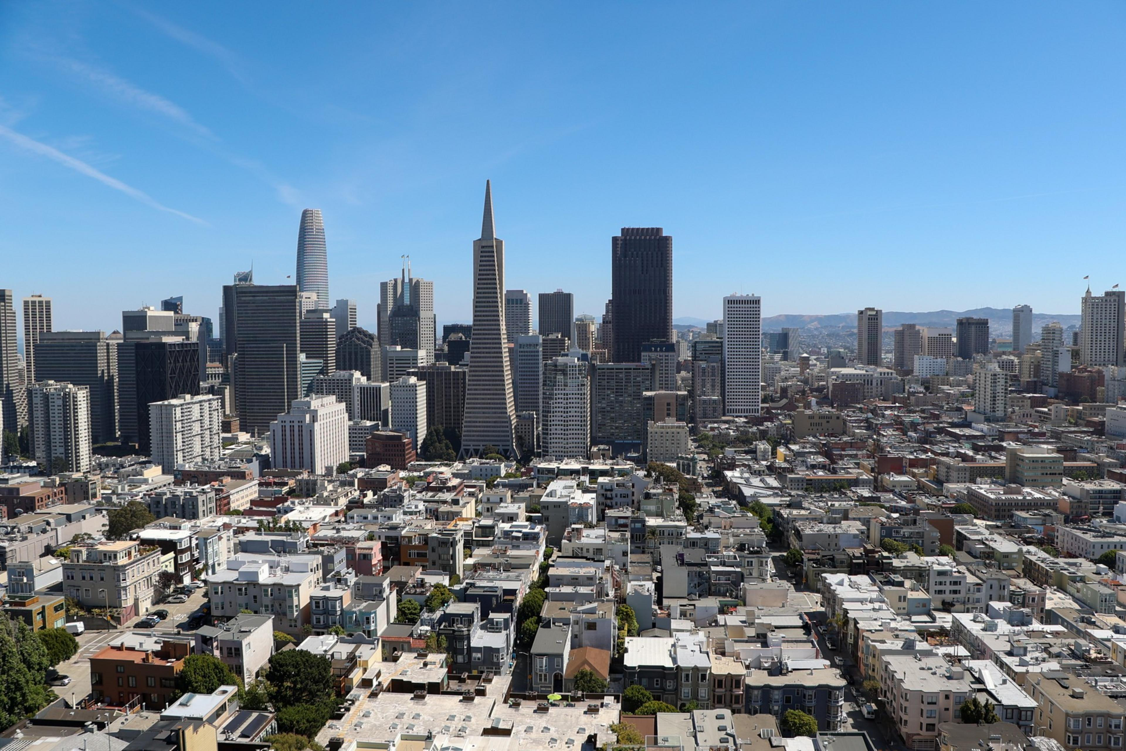 A city skyline with a mix of tall skyscrapers and shorter buildings under a clear blue sky, with streets and houses visible in the foreground.