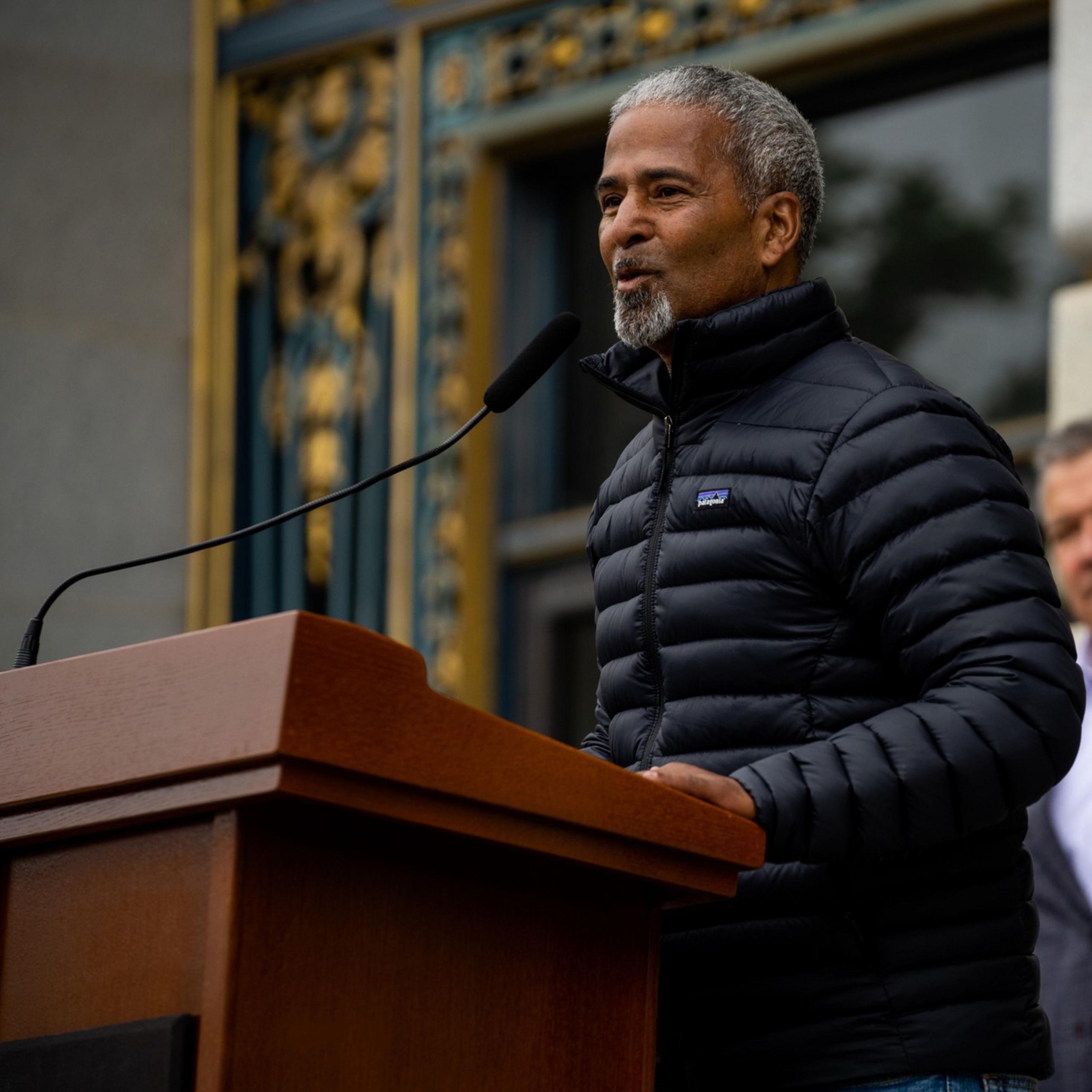 A man in a black puffer jacket speaks at a podium with a microphone. Another man stands in the background, wearing a blazer. The setting appears formal.