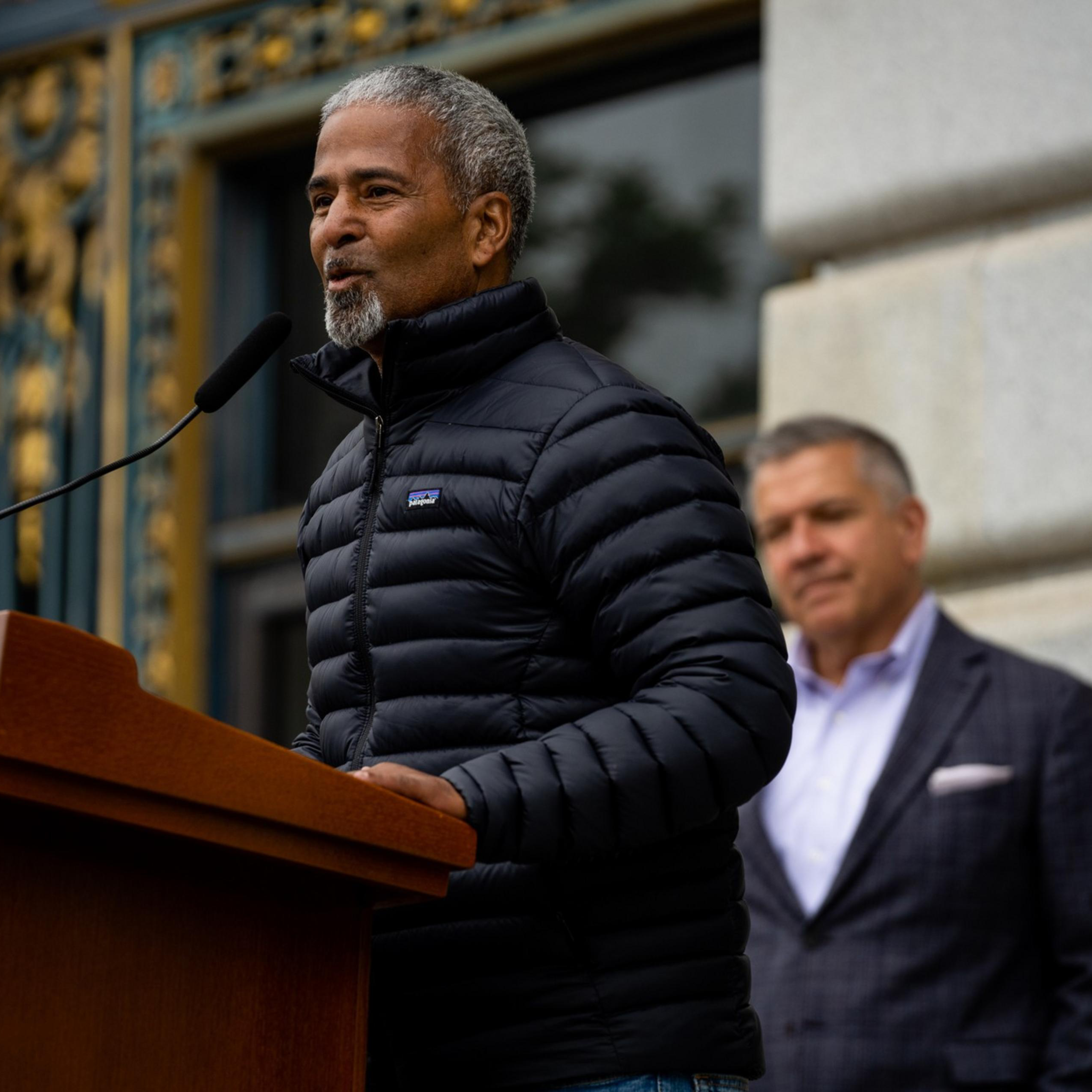 A man in a black puffer jacket speaks at a podium with a microphone. Another man stands in the background, wearing a blazer. The setting appears formal.