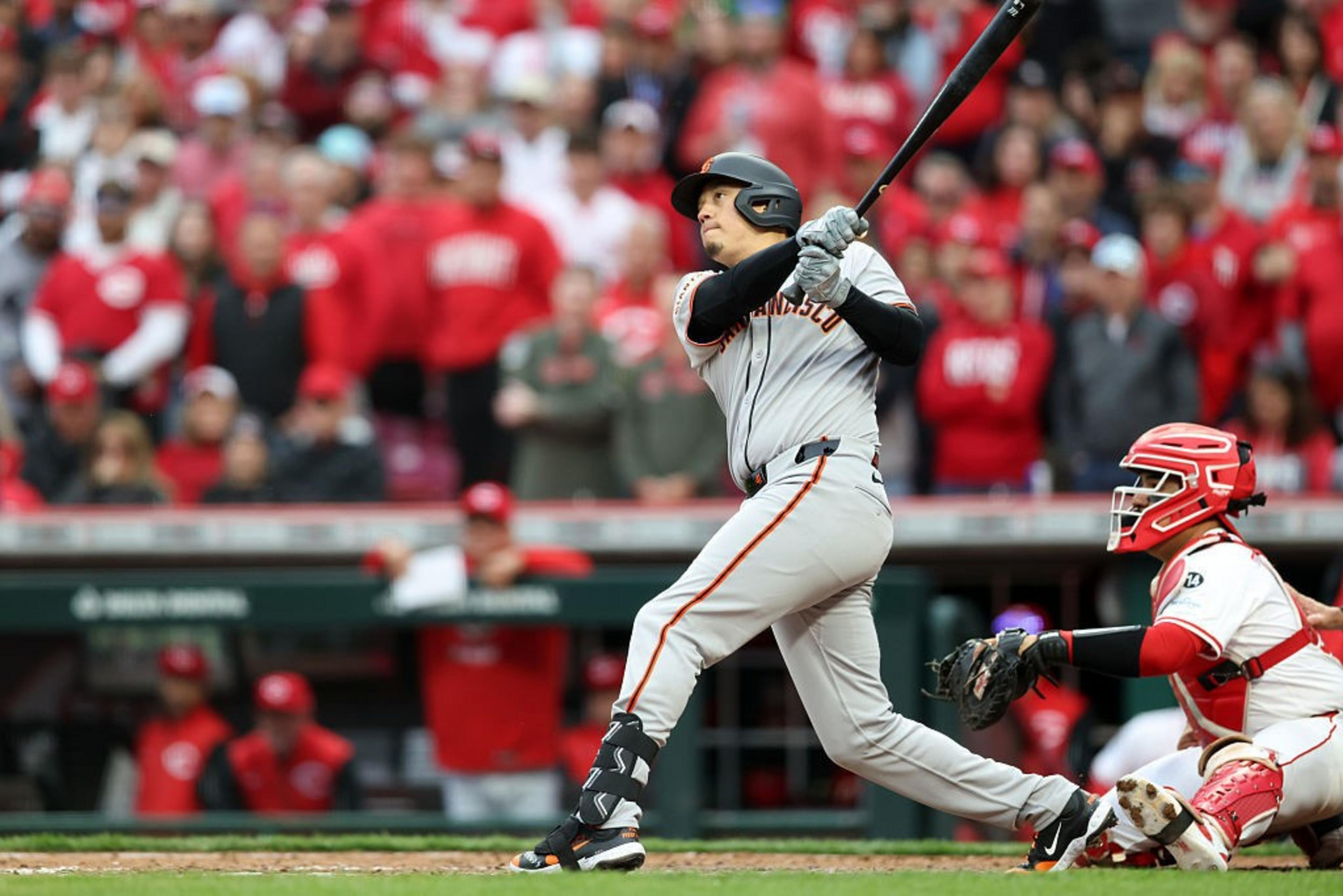 A baseball player in a gray uniform swings his bat during a game. A catcher in red gear crouches behind him. The crowd in the background is mostly in red.