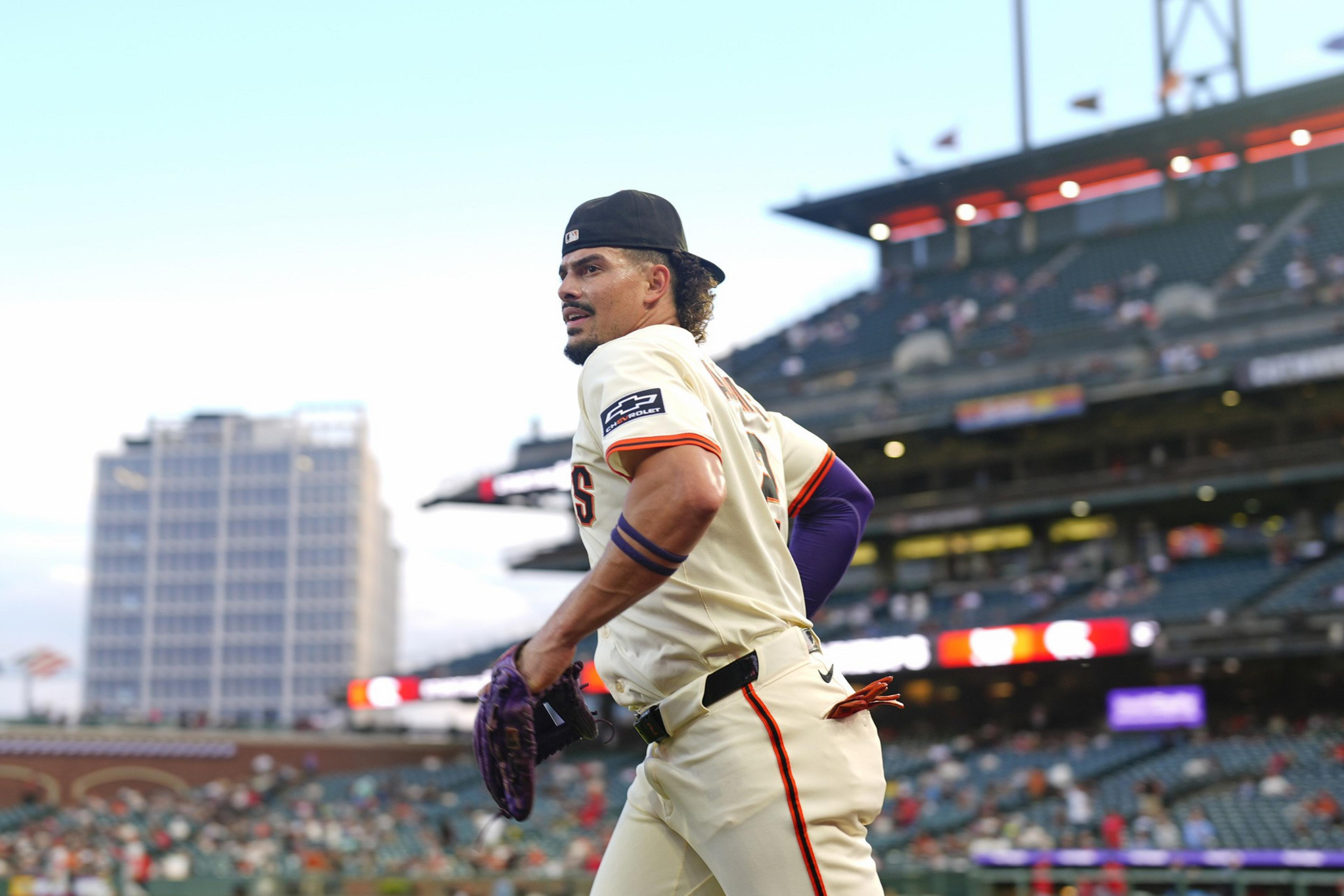 A baseball player in a cream uniform and backward cap walks on the field with a glove, stadium seats and a building visible in the background.