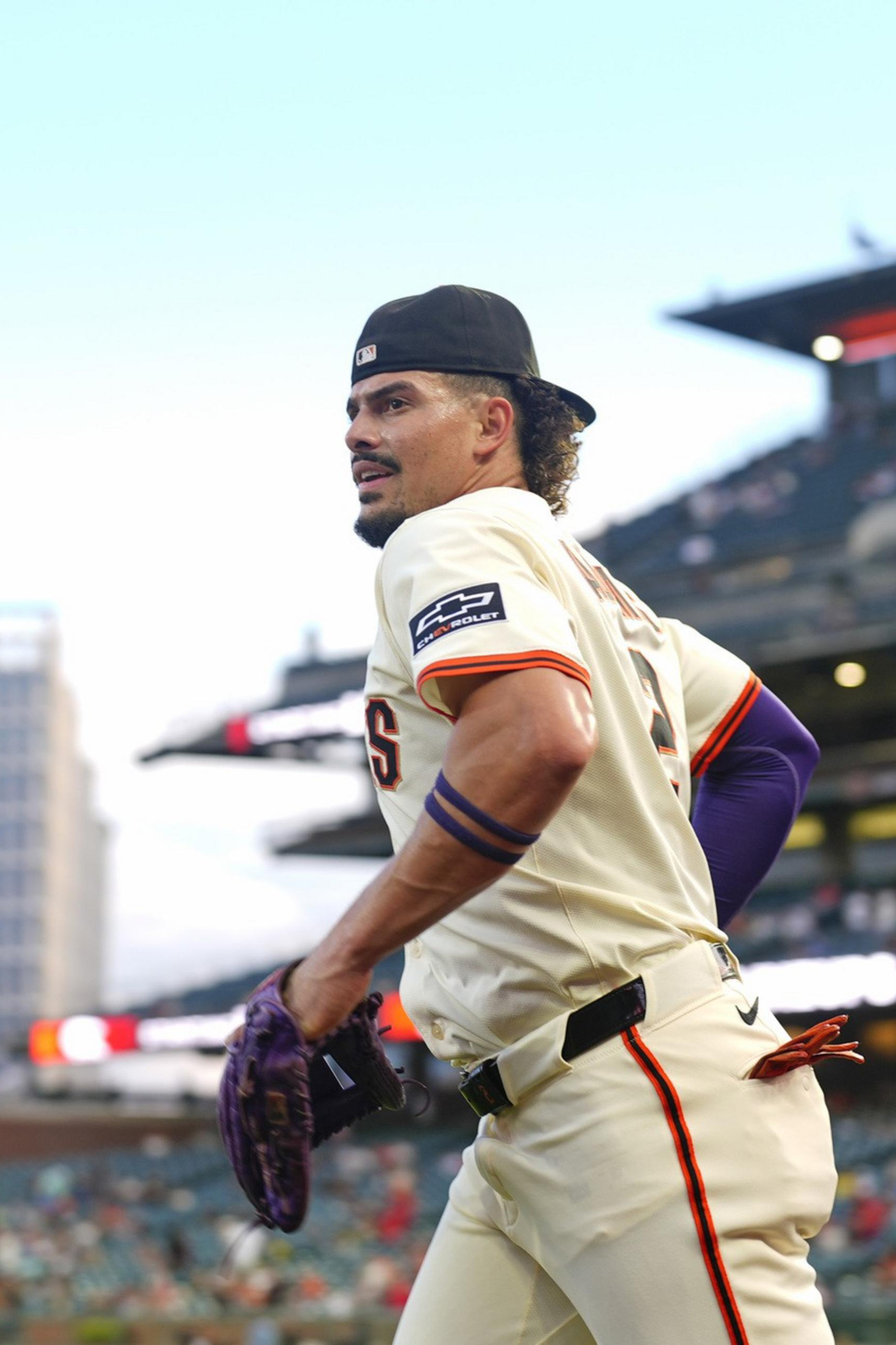 A baseball player in a cream uniform and backward cap walks on the field with a glove, stadium seats and a building visible in the background.