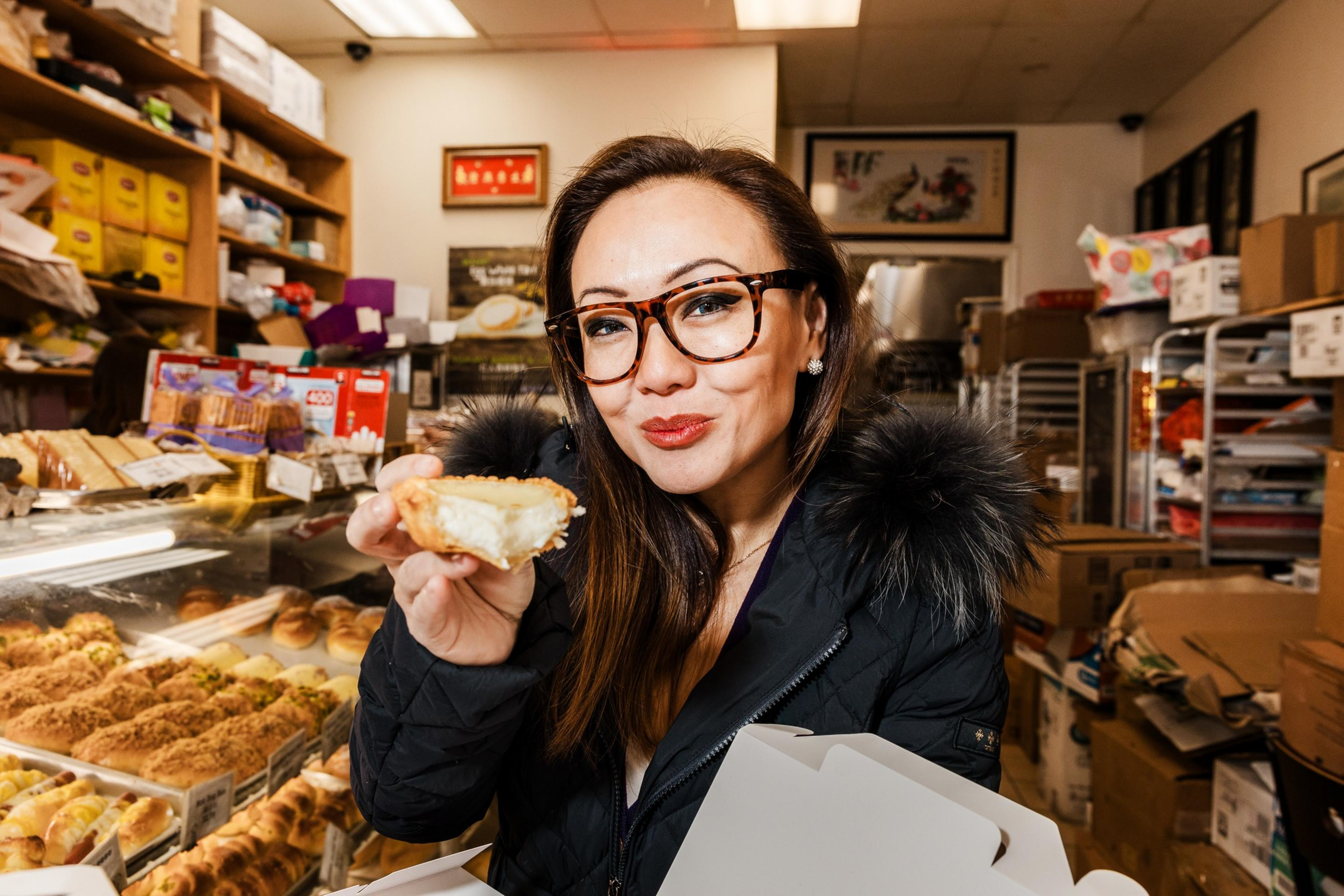 A person in a warm coat and glasses is inside a bakery, smiling while holding a partially eaten pastry. The background shows shelves filled with baked goods.