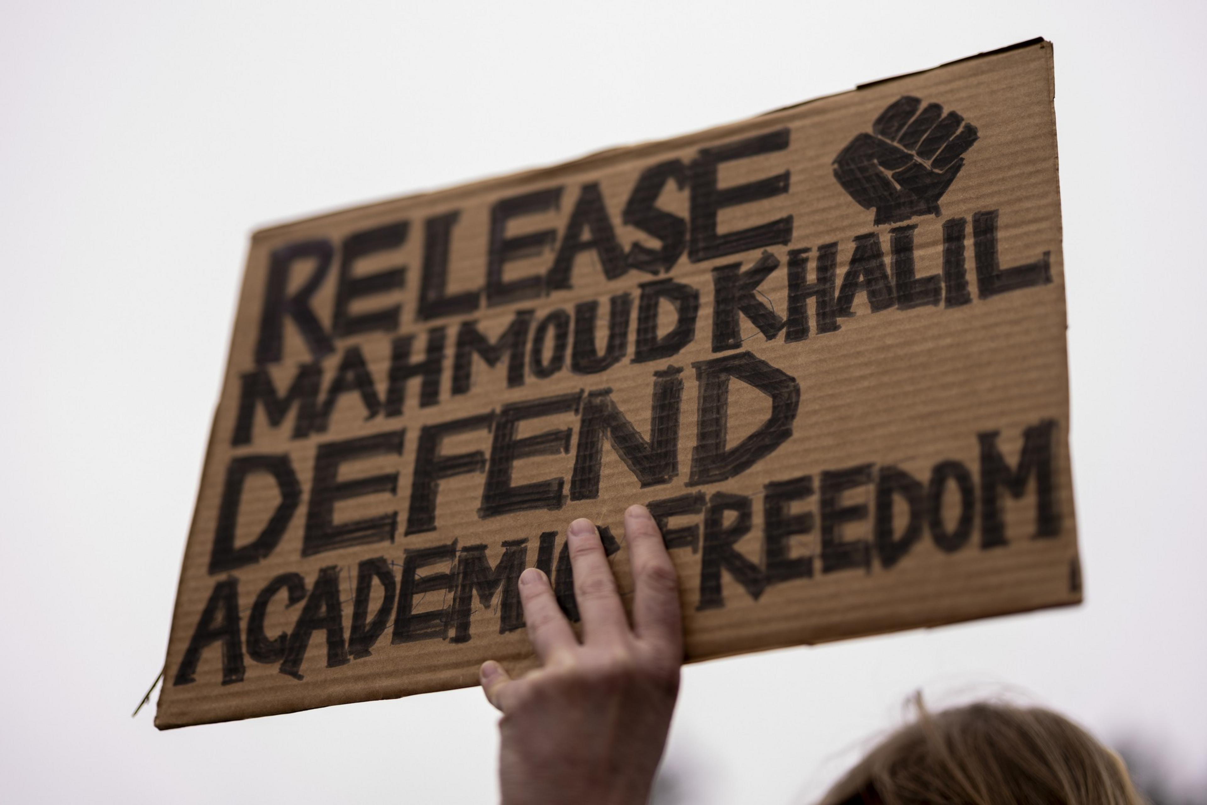 A hand holds a cardboard sign with bold text reading &quot;Release Mahmoud Khalil, Defend Academic Freedom&quot; alongside a drawn fist.