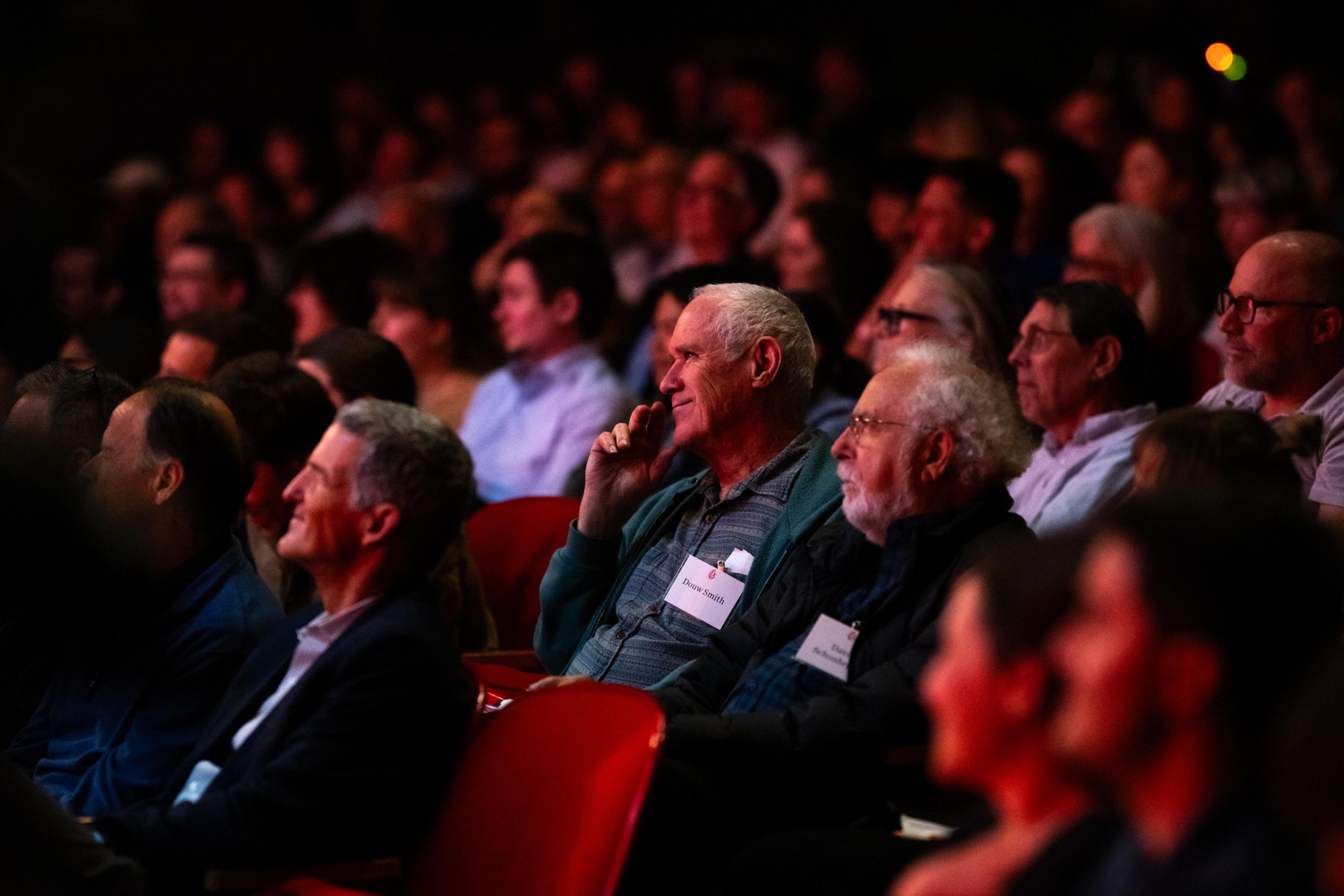 A large group of people, mostly older men, are seated in a dark auditorium, attentively listening. They wear name tags and appear engaged in the event.