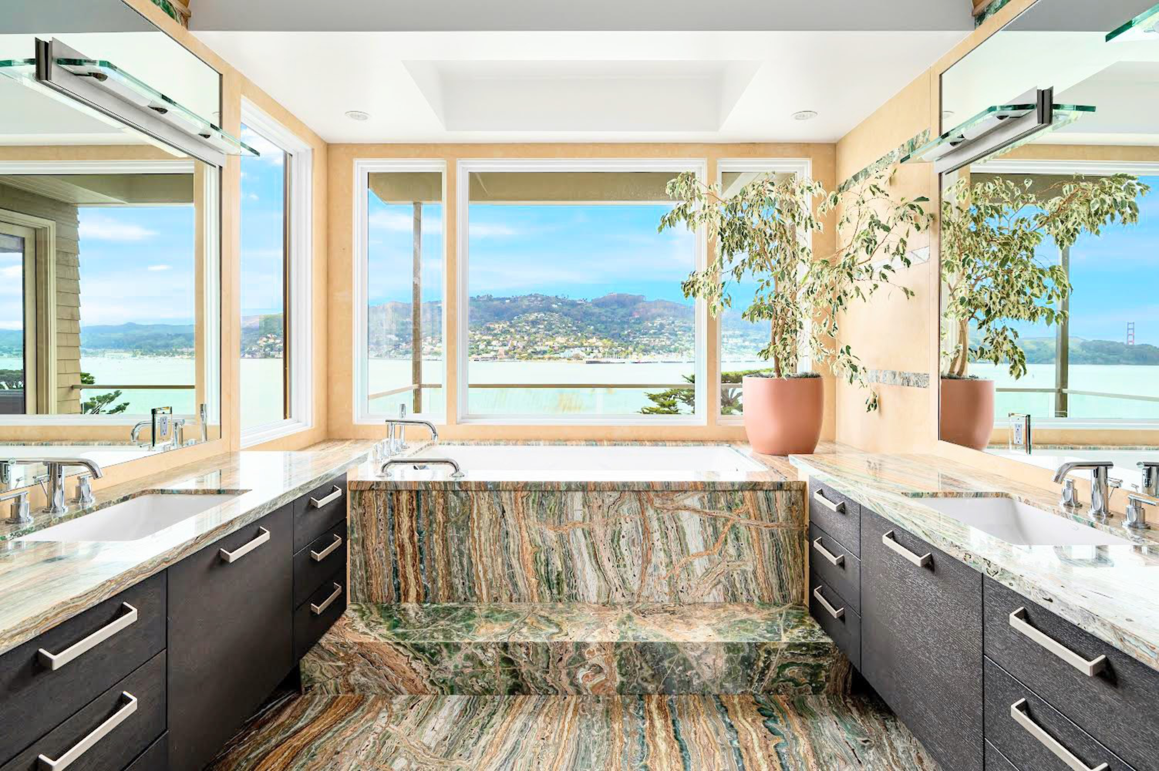 A luxurious bathroom features a large marble tub with steps, dual sinks with dark cabinetry, and large windows showing a scenic water and hill view.