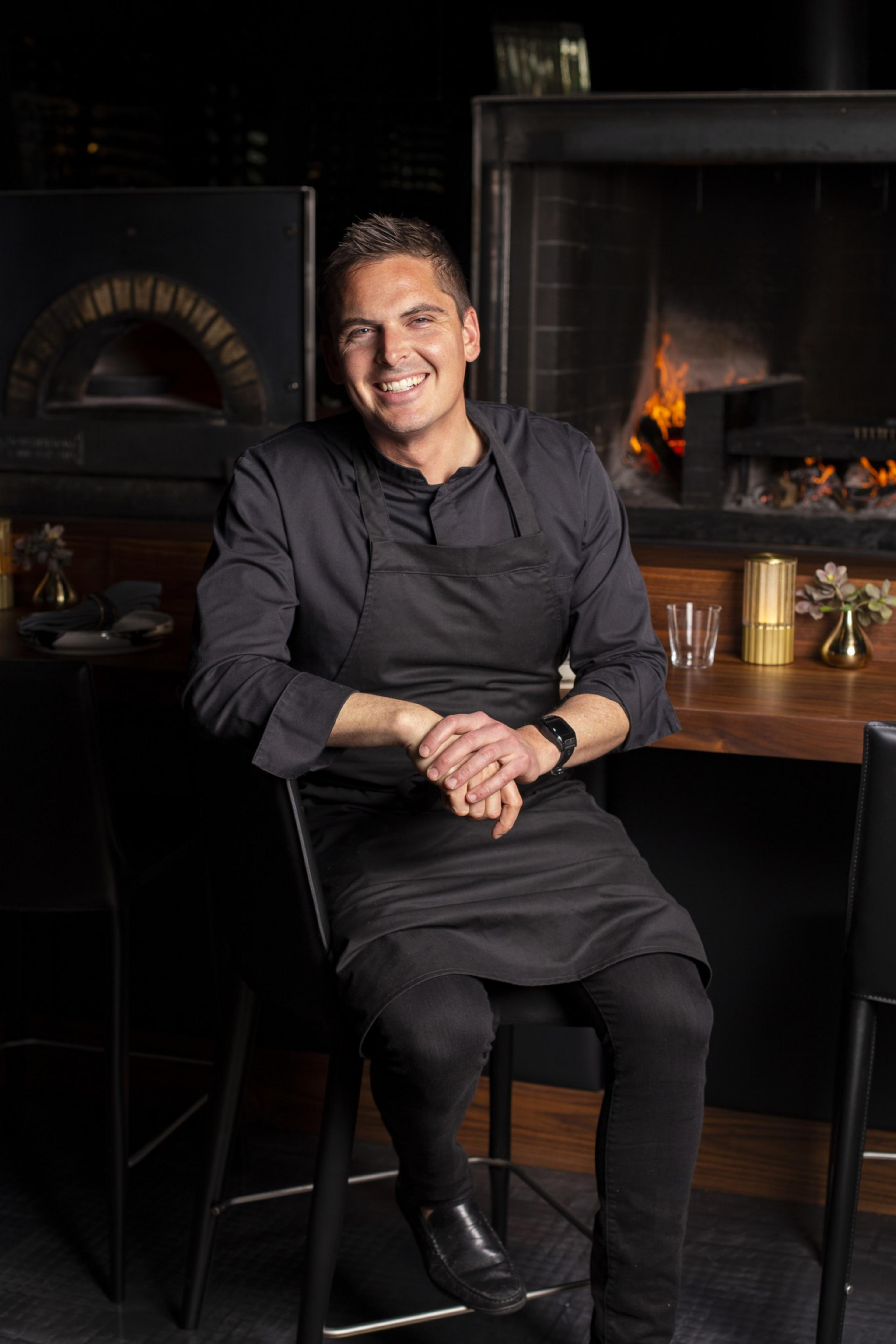 A smiling person in a black chef's outfit sits on a chair in a warmly lit kitchen, with a wood-fired oven and fireplace in the background.