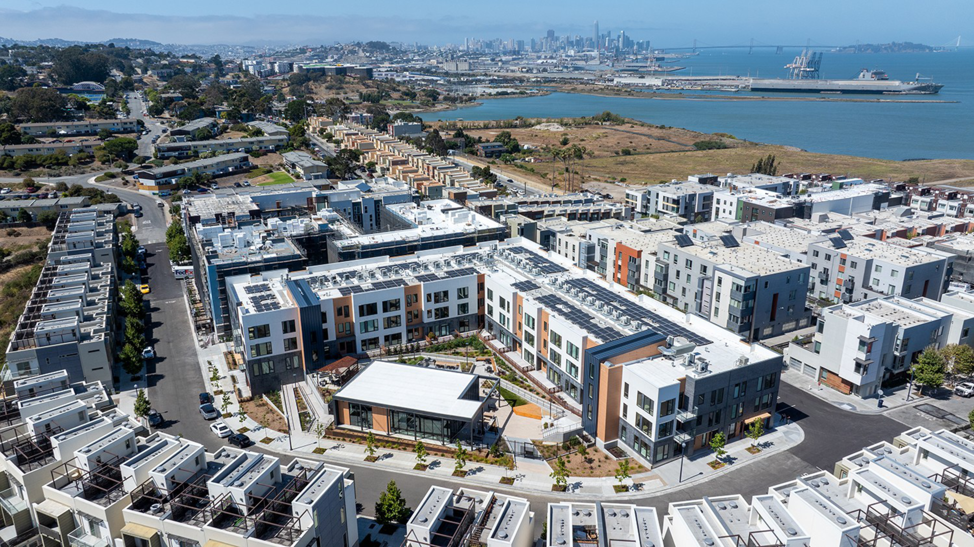 A modern residential complex with multiple white and gray buildings surrounds a central courtyard, with a bay and distant city skyline in the background.