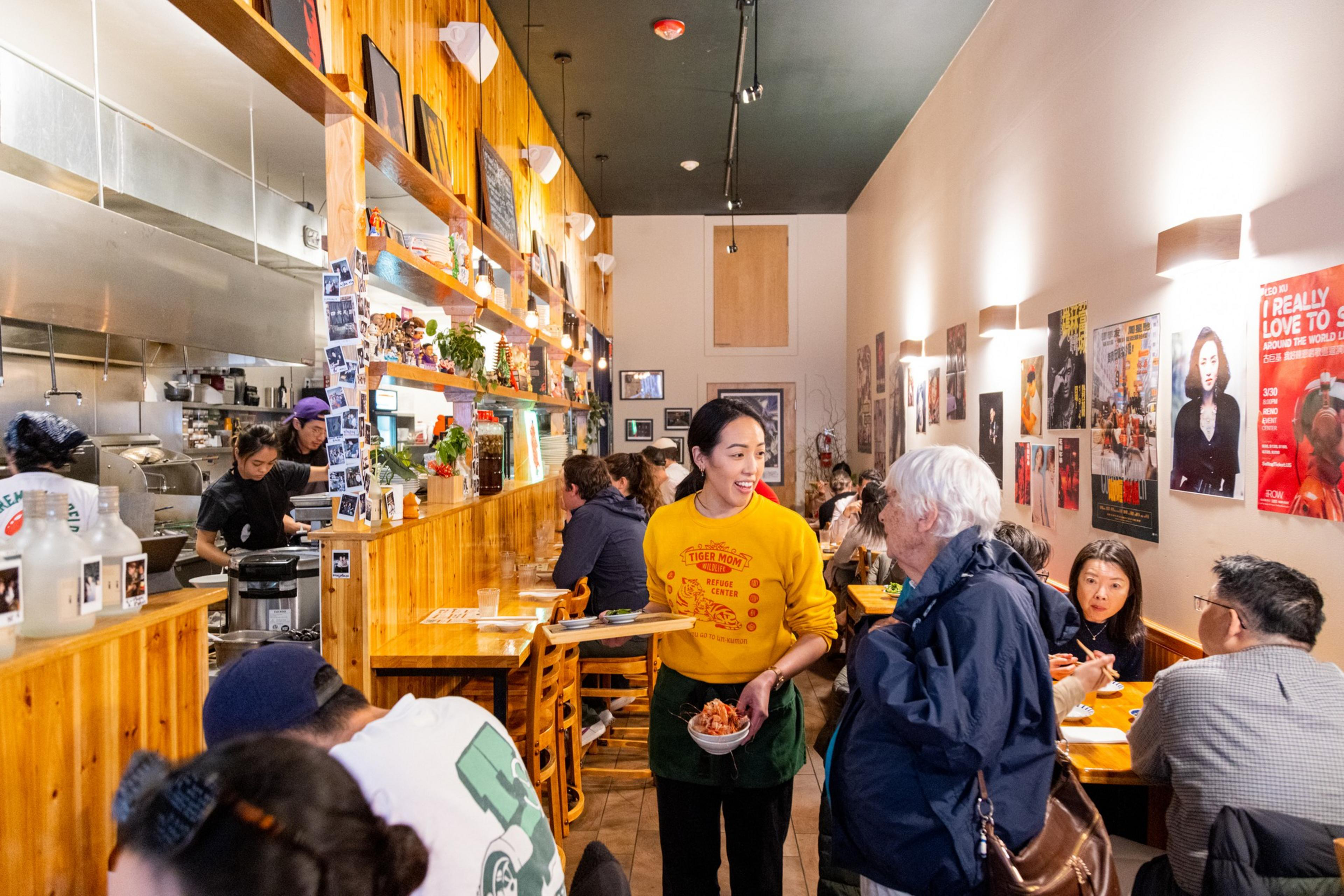 The image shows a busy restaurant with wooden decor. A server in a yellow shirt talks to a customer, while diners eat at closely packed tables. Walls have colorful posters.