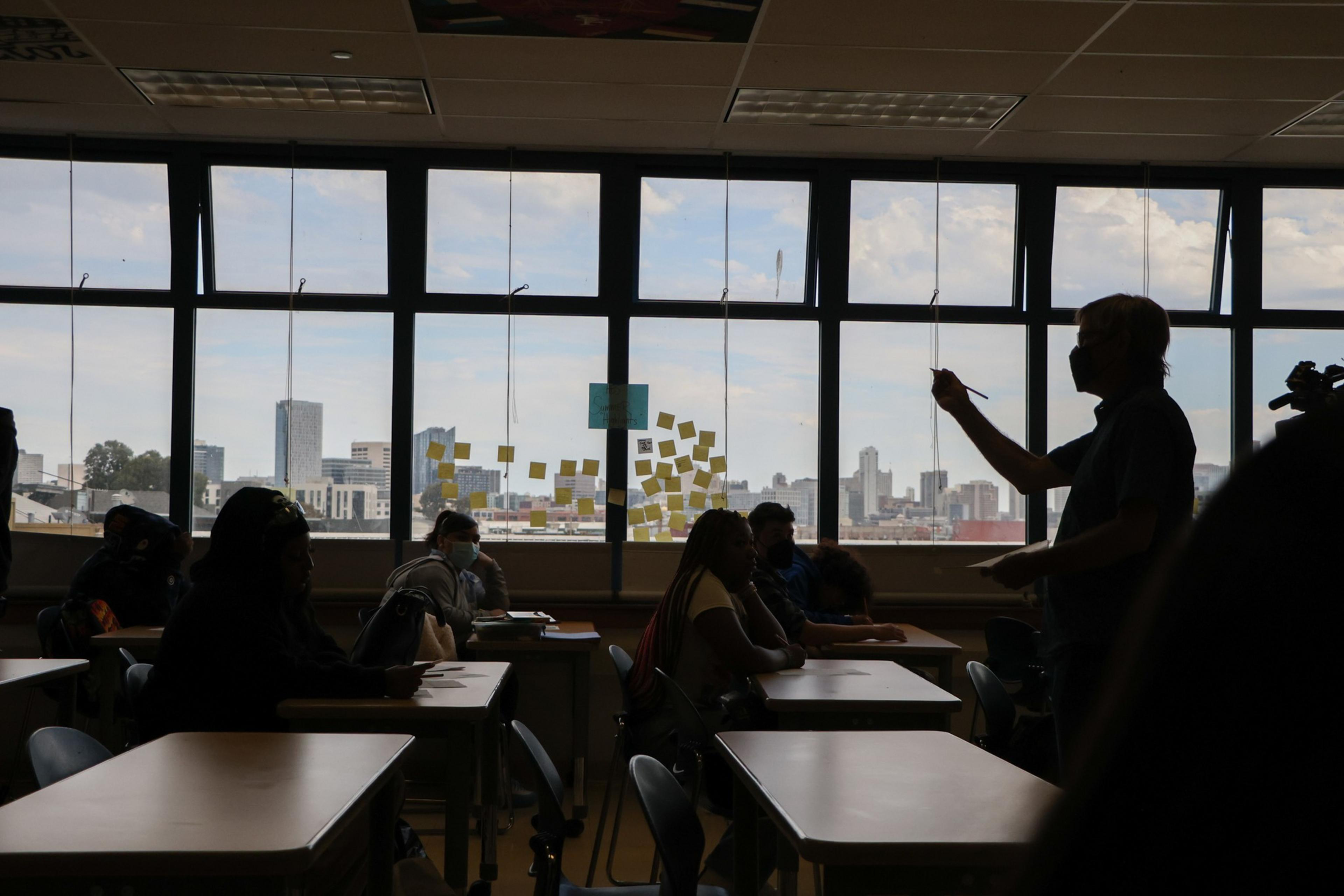 A teacher wearing a mask stands in a classroom pointing at seated students. Large windows behind them reveal a city skyline and post-it notes on the glass.