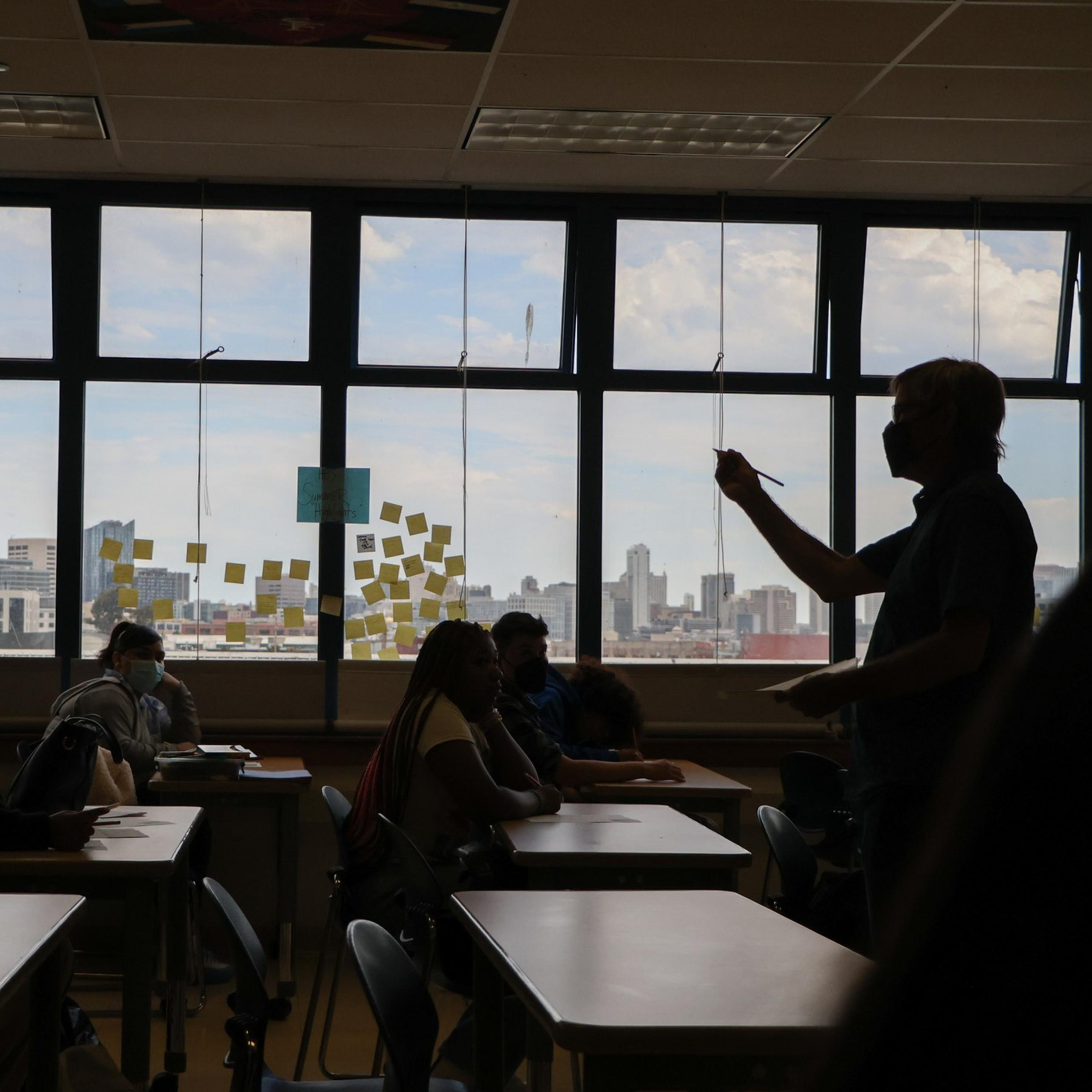 A teacher wearing a mask stands in a classroom pointing at seated students. Large windows behind them reveal a city skyline and post-it notes on the glass.