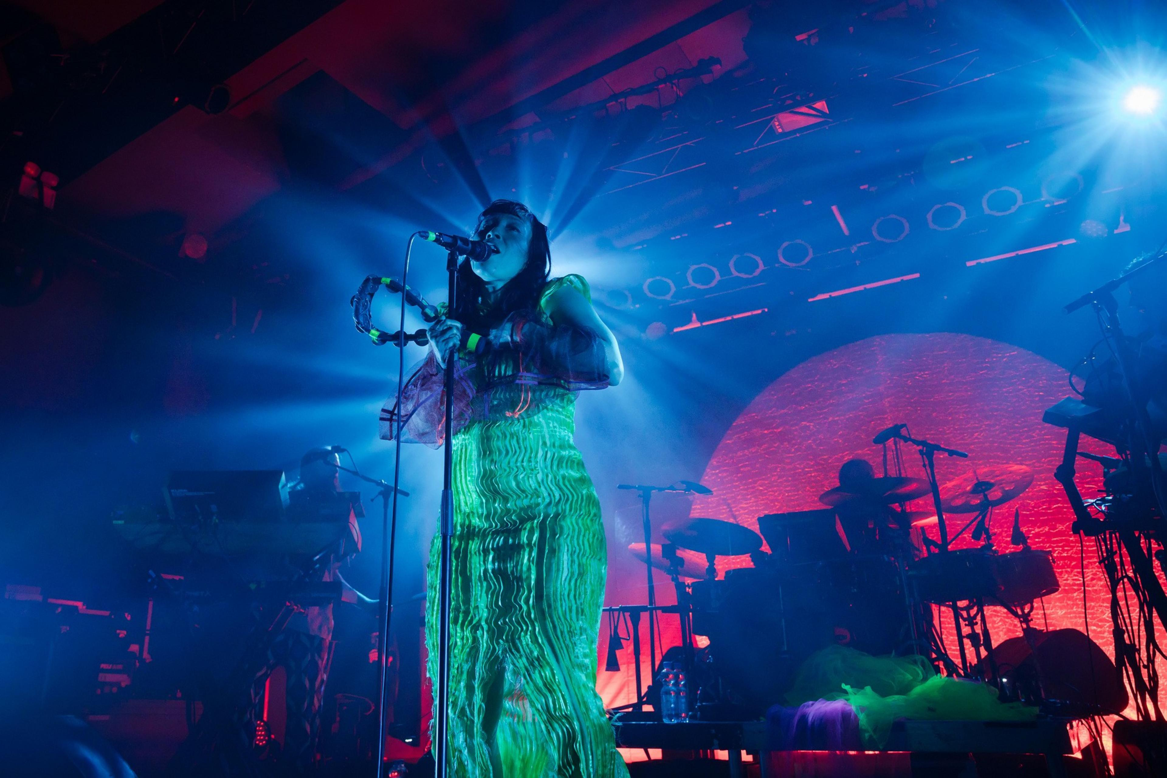 A singer in a green dress on a dark festival stage is lit from behind and shot at a low angle