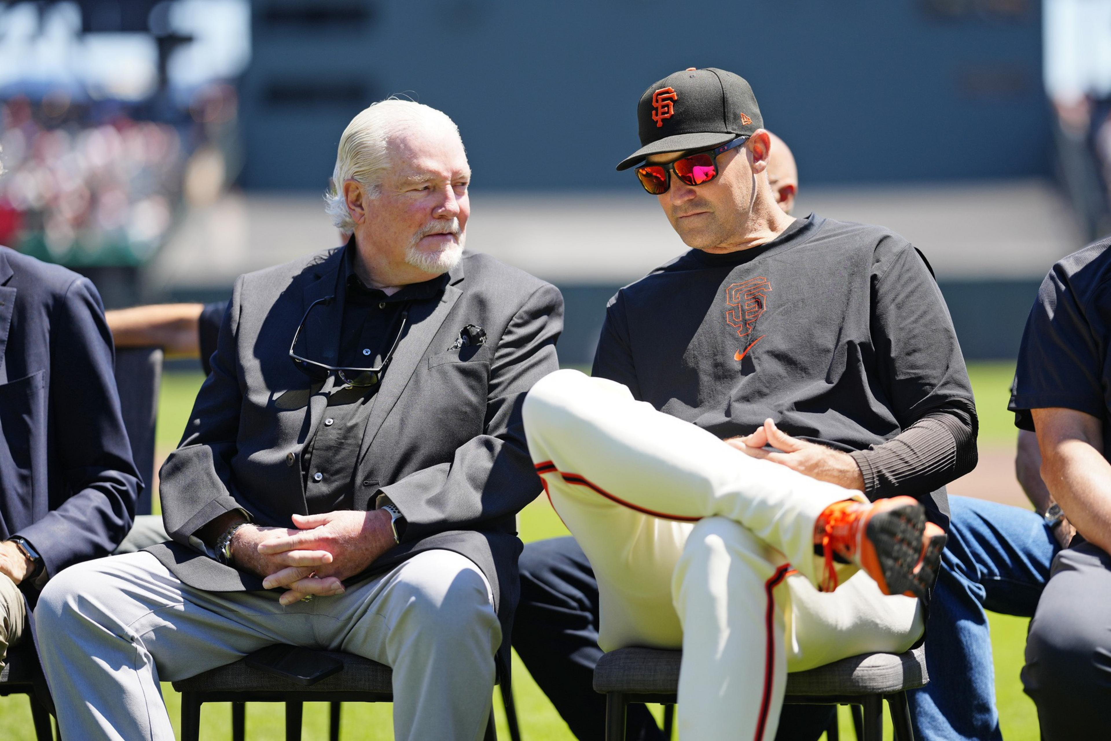 Two men sit outdoors. One wears a black suit with sunglasses on his chest, the other wears a baseball cap and jersey with &quot;SF&quot; logo, and white pants.