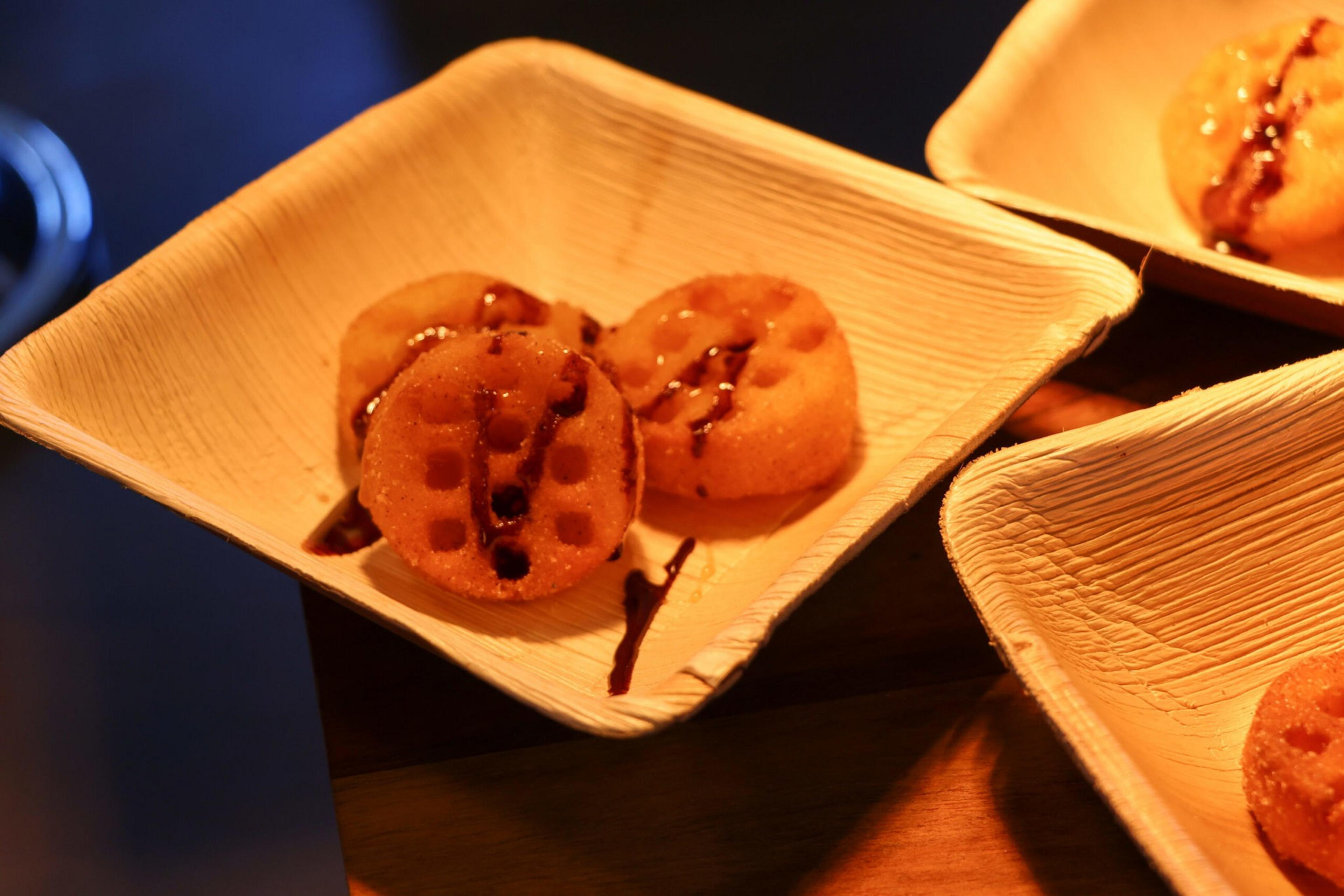 Three round, waffle-textured sweets drizzled with chocolate sauce sit on a square, natural-fiber plate under warm lighting.