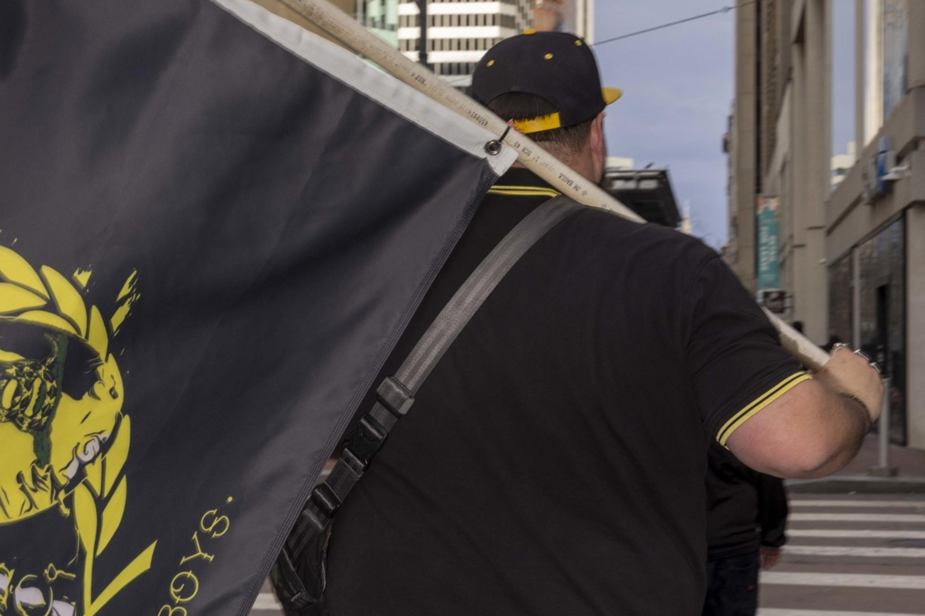 A person is walking down a city street, carrying a large black flag with yellow designs and text. They're wearing jeans and a black cap, surrounded by tall buildings.
