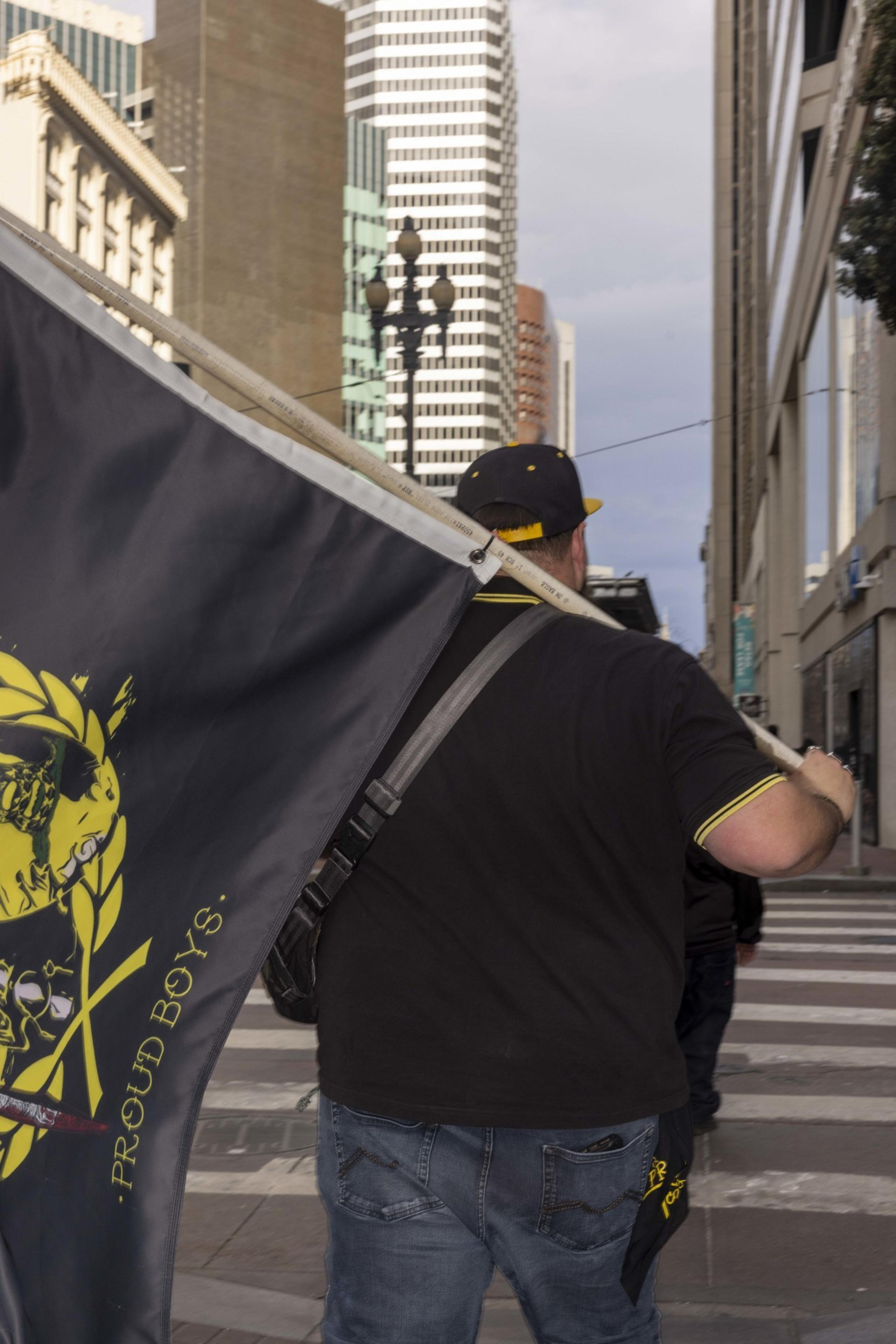 A person is walking down a city street, carrying a large black flag with yellow designs and text. They're wearing jeans and a black cap, surrounded by tall buildings.