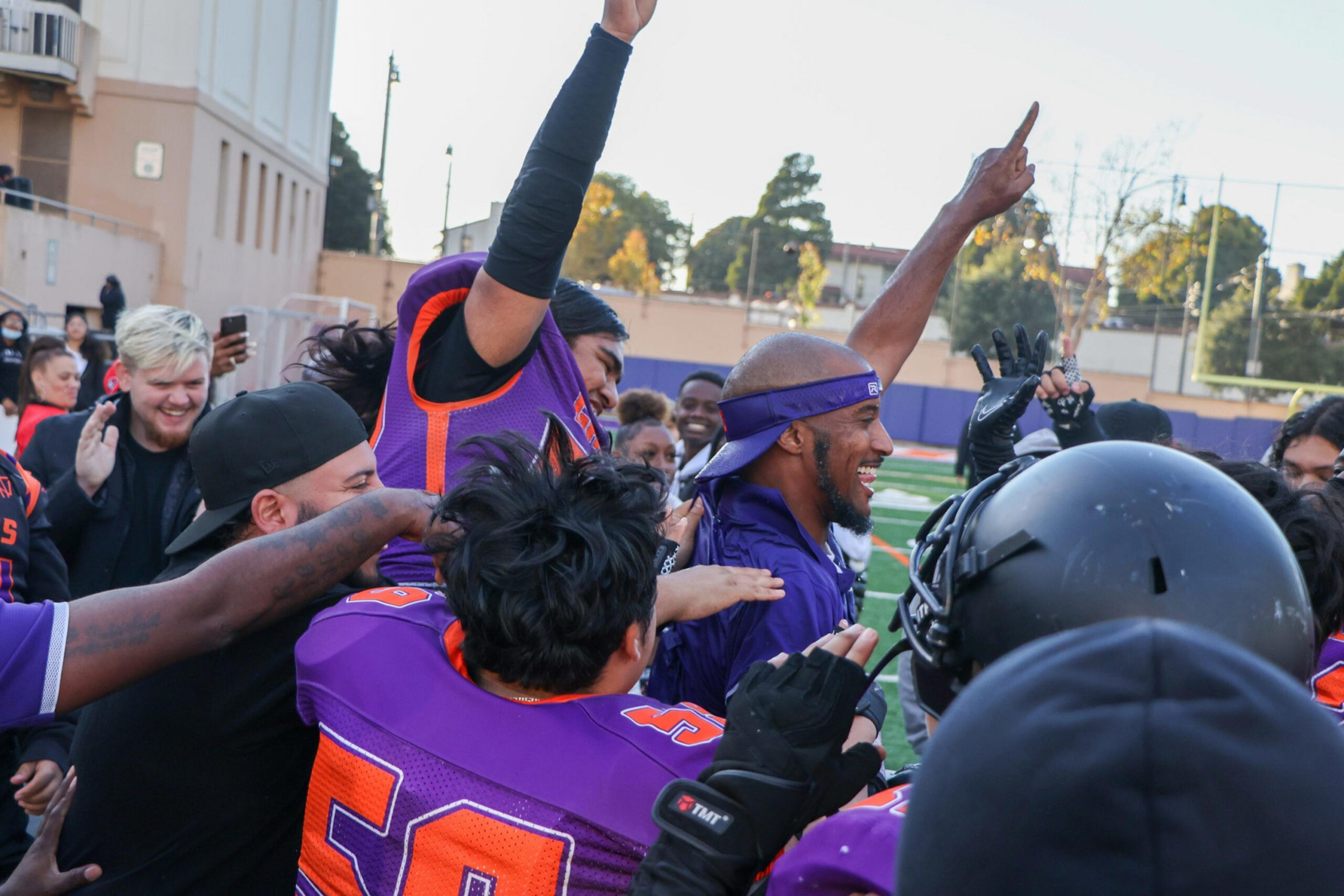 A group of football players and coaches in purple and orange uniforms celebrate energetically on a field, raising hands and smiling with excitement.