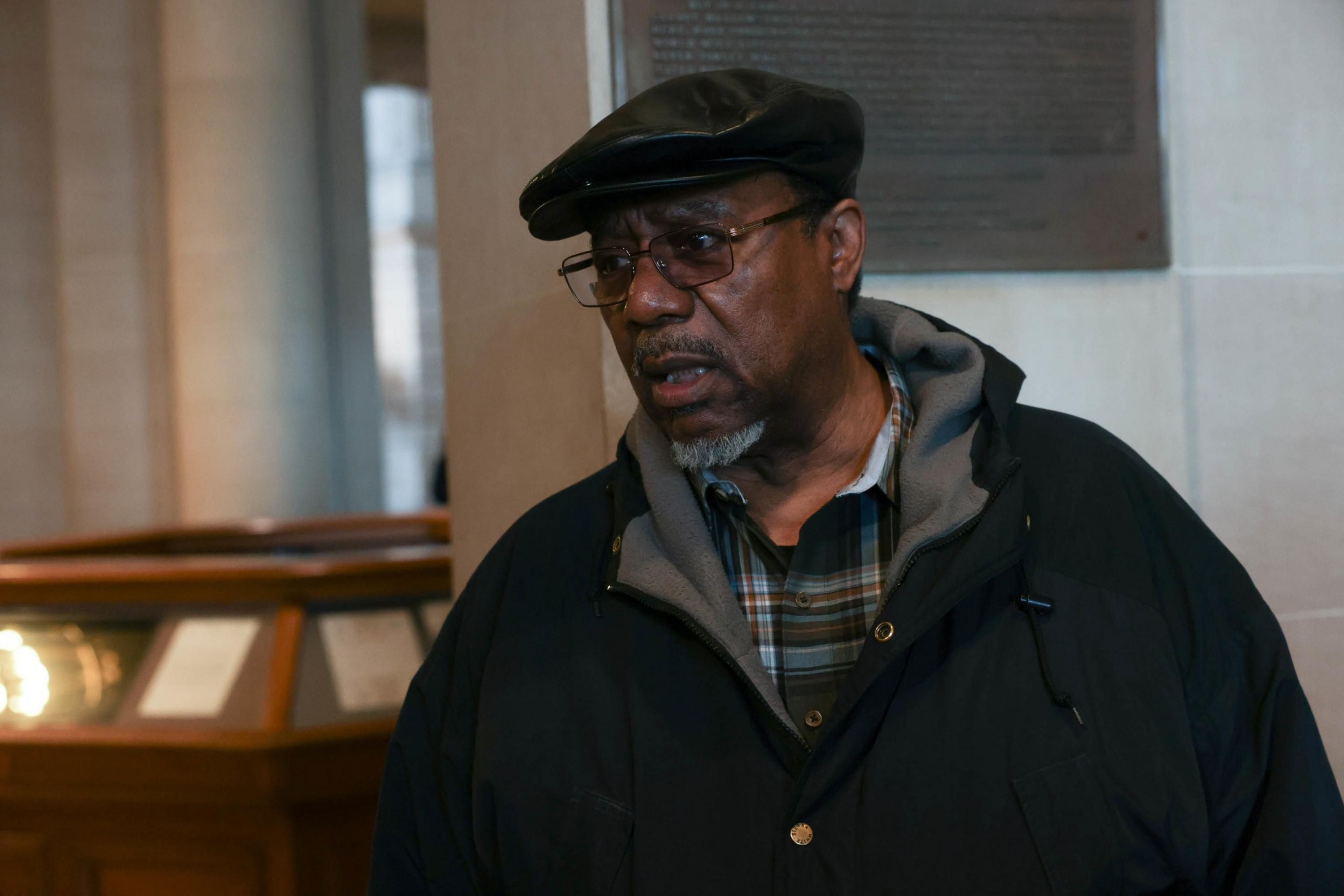 An older man with glasses and a cap stands indoors, wearing a jacket over a plaid shirt.