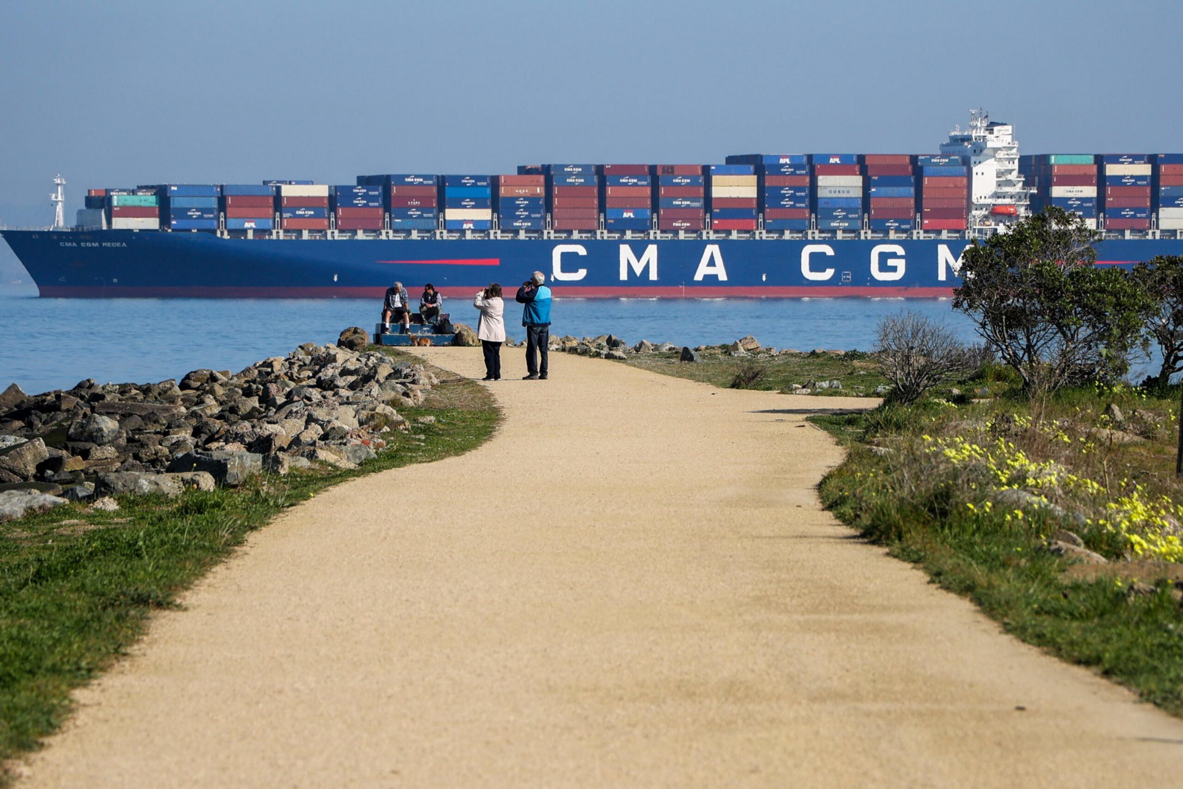 A large container ship loaded with colorful shipping containers sails near a curved walking path where people are standing and sitting.