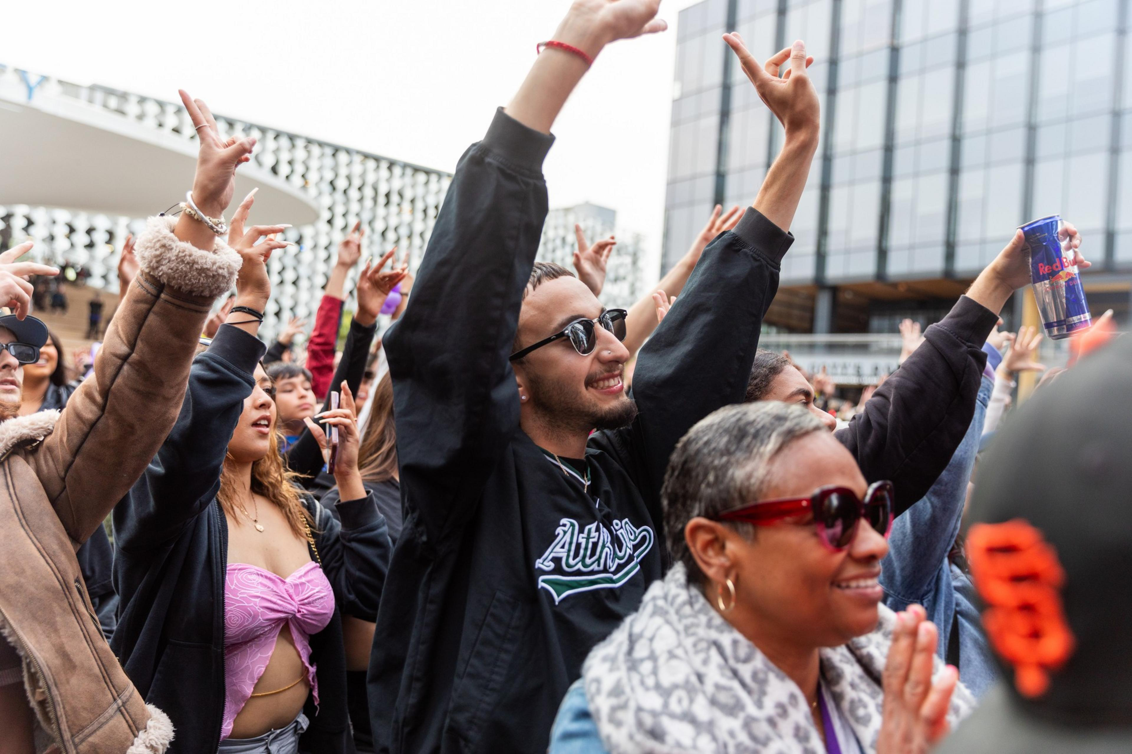 A diverse group of people enjoying a public event, raising their hands, smiling, and holding drinks, expressing excitement and happiness.