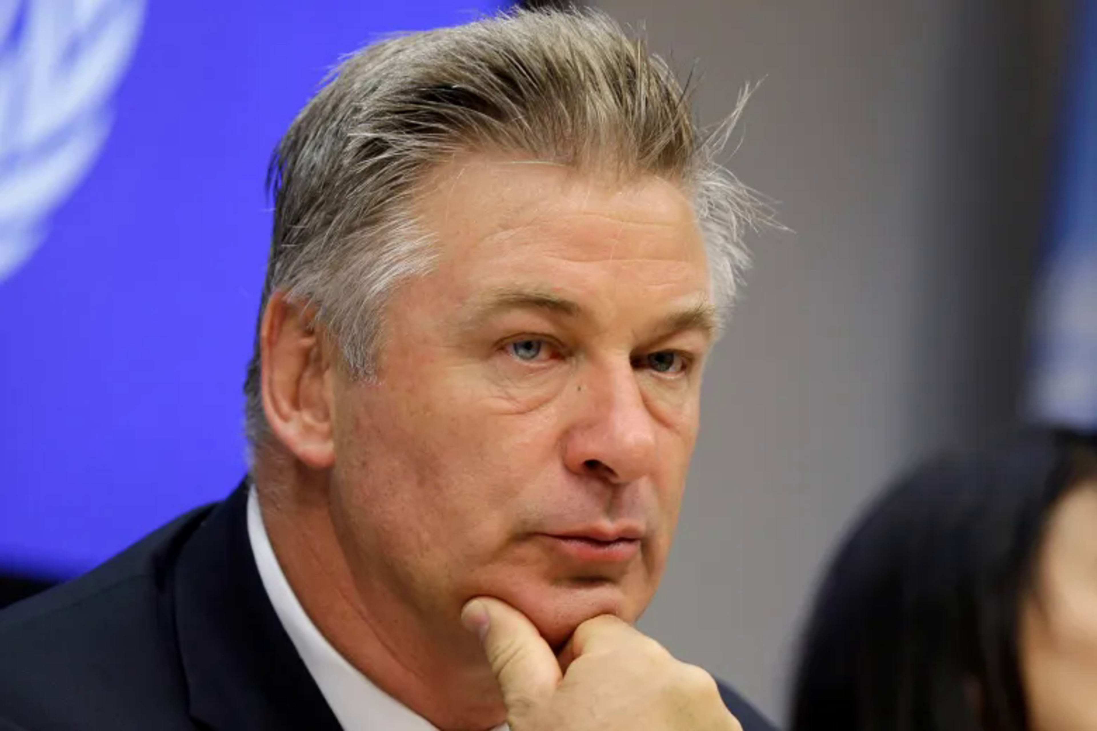 A middle-aged man with grayish hair rests his chin on his hand, looking thoughtful against a blue and gray blurred background.