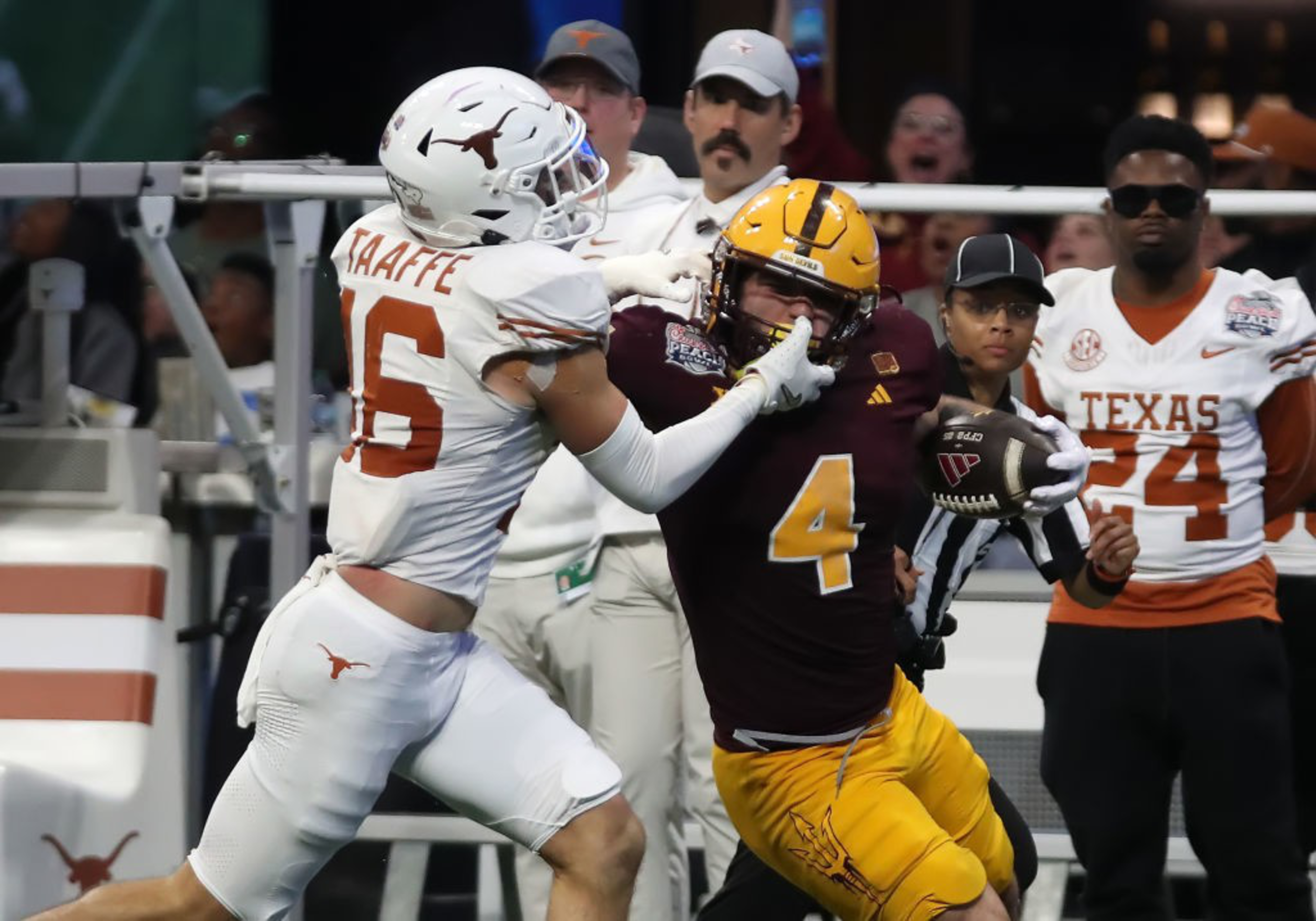 A football player in white tackles a player in dark maroon and yellow, who is carrying the ball, while a referee and others watch intently from the sidelines.