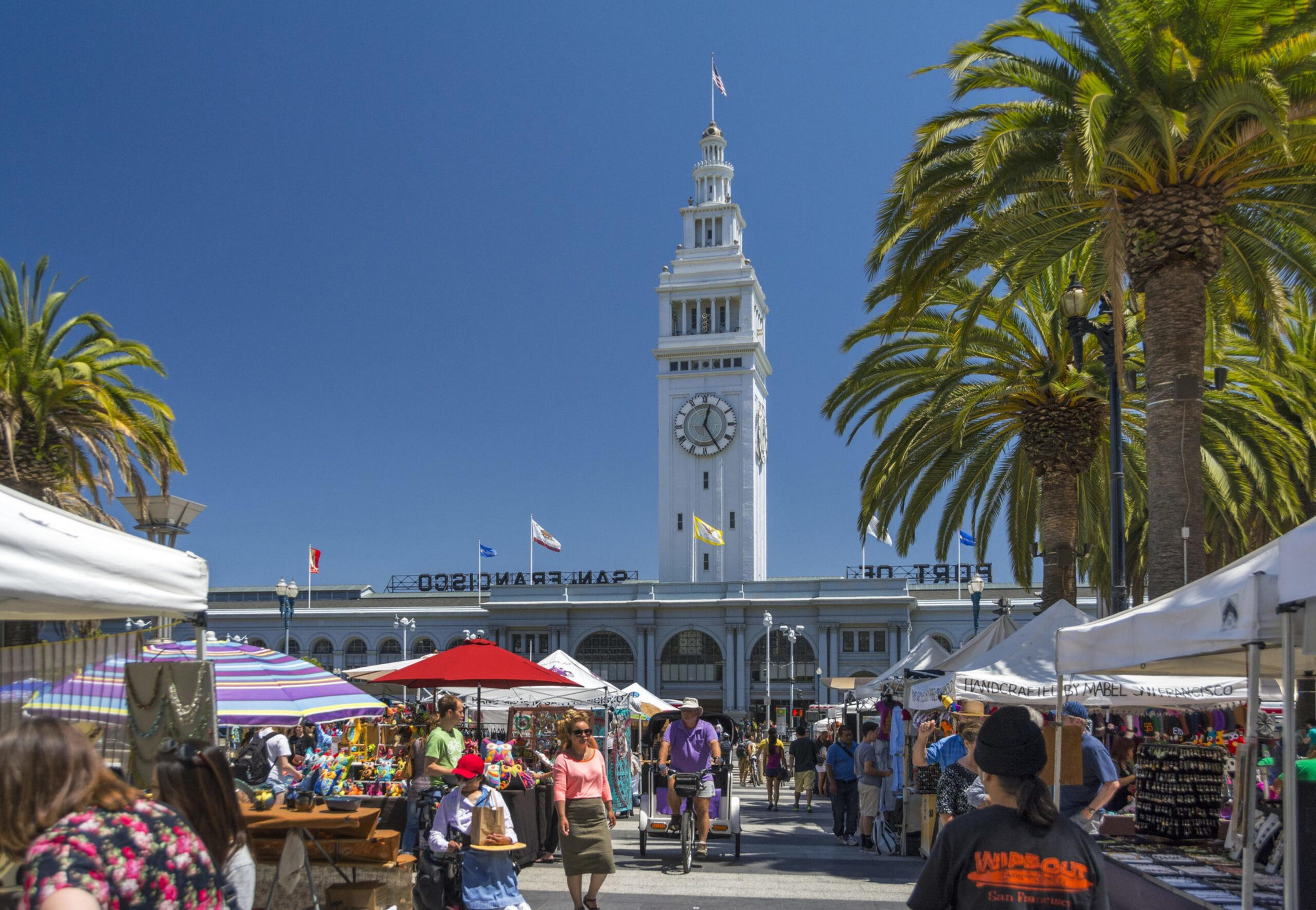 A busy outdoor market with colorful stalls and umbrellas is set in front of a tall clock tower labeled "San Francisco" under a clear blue sky.
