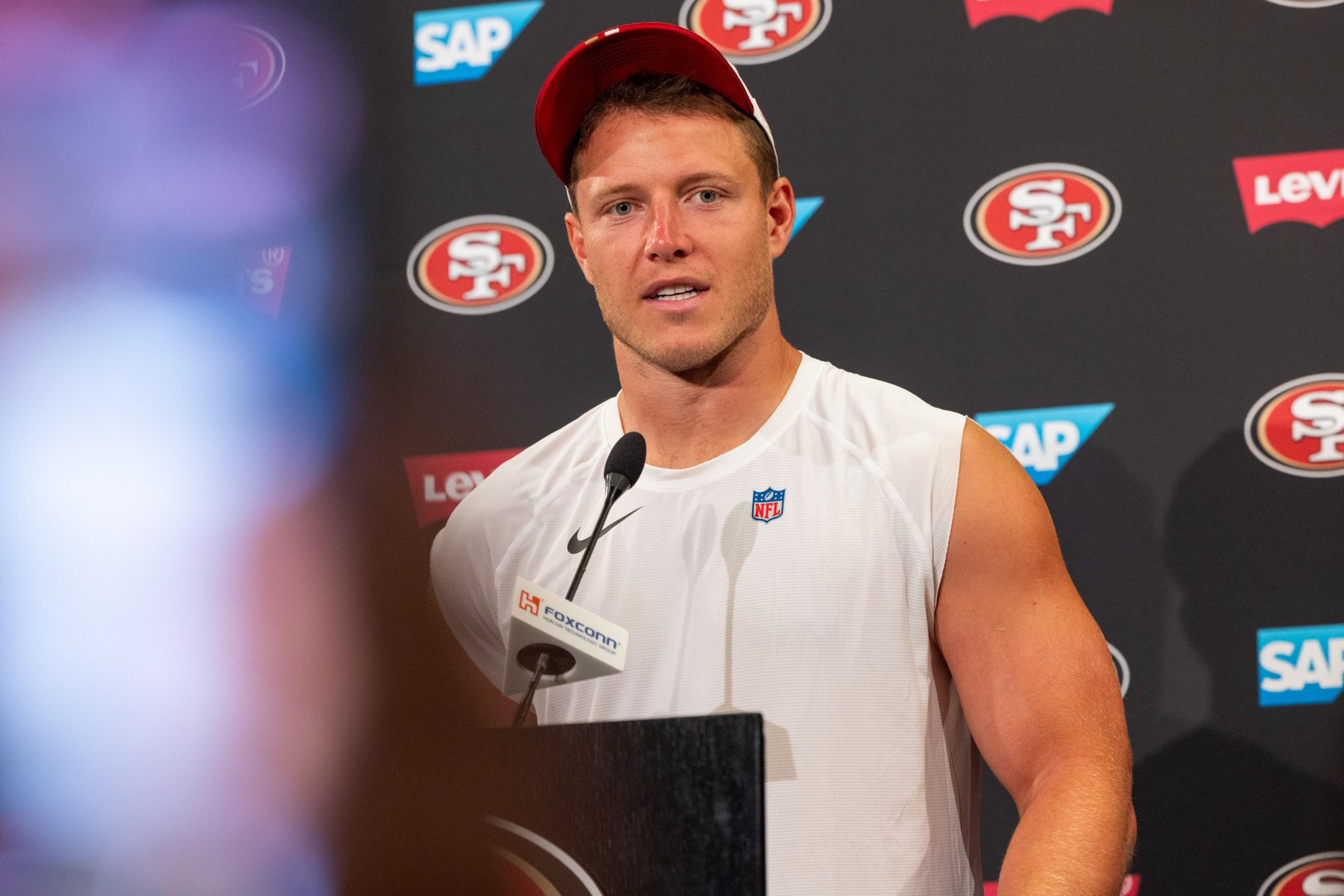 A muscular man in a white sleeveless NFL shirt and red cap speaks at a podium with San Francisco 49ers logos and sponsor signs in the background.