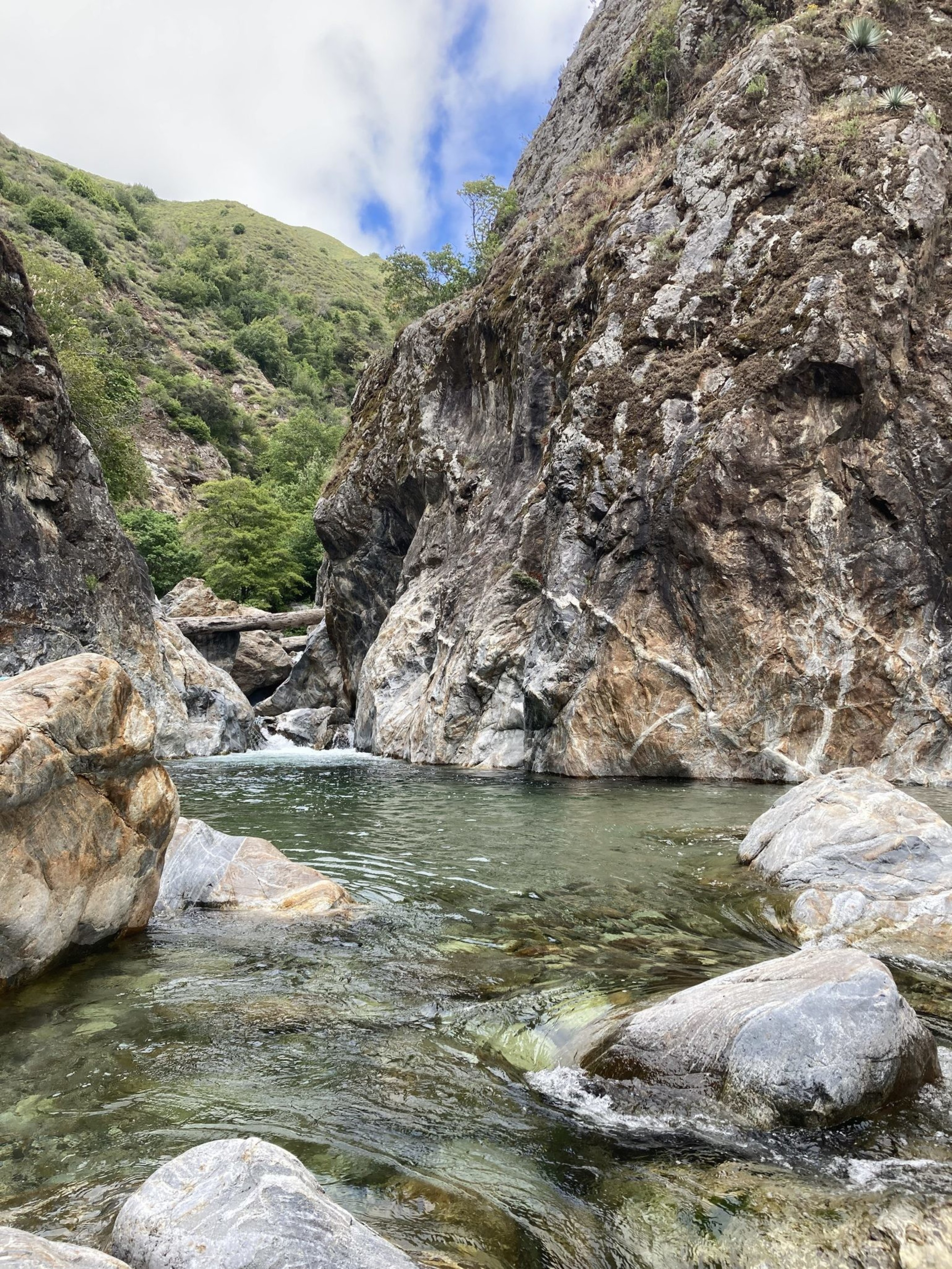 The image shows a clear river flowing between large rocky cliffs with some greenery on them, surrounded by lush green hills under a partly cloudy sky.