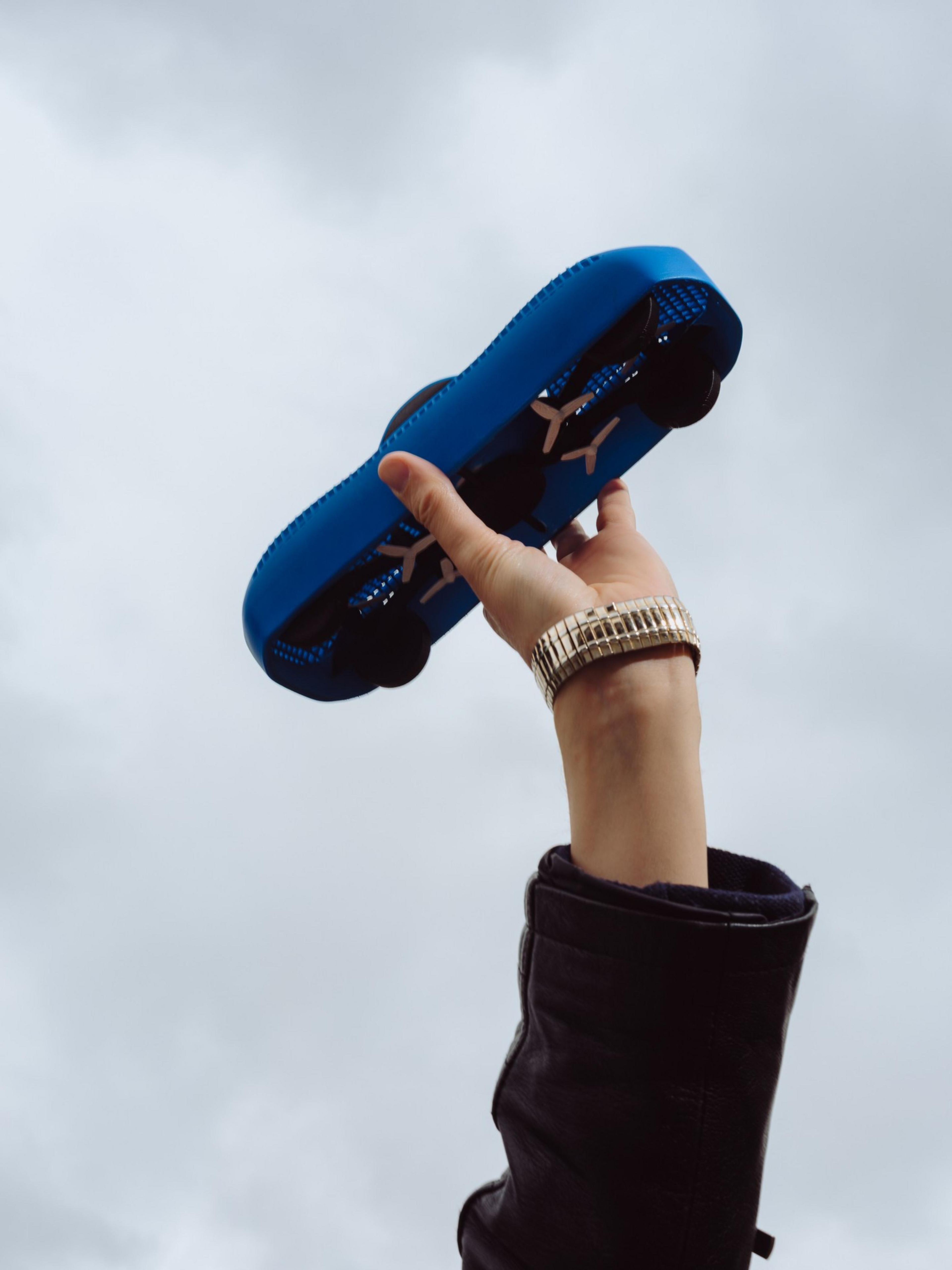 A hand in a black sleeve and a gold watch holds up a blue futuristic shoe with visible propellers against a cloudy sky.