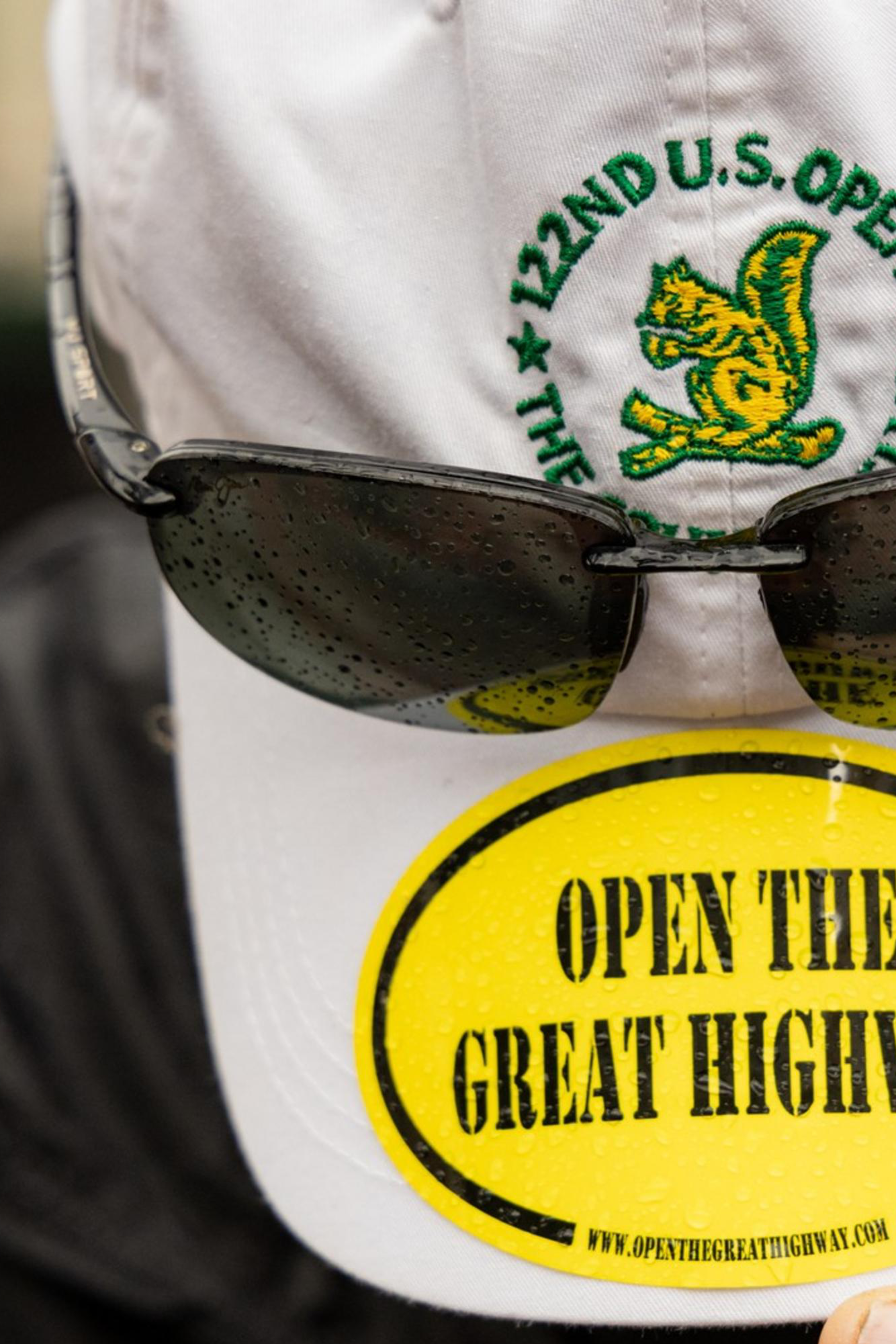 A person in a black jacket holds the brim of a white cap with dark sunglasses attached. The cap displays a yellow sticker reading &quot;OPEN THE GREAT HIGHWAY.&quot;