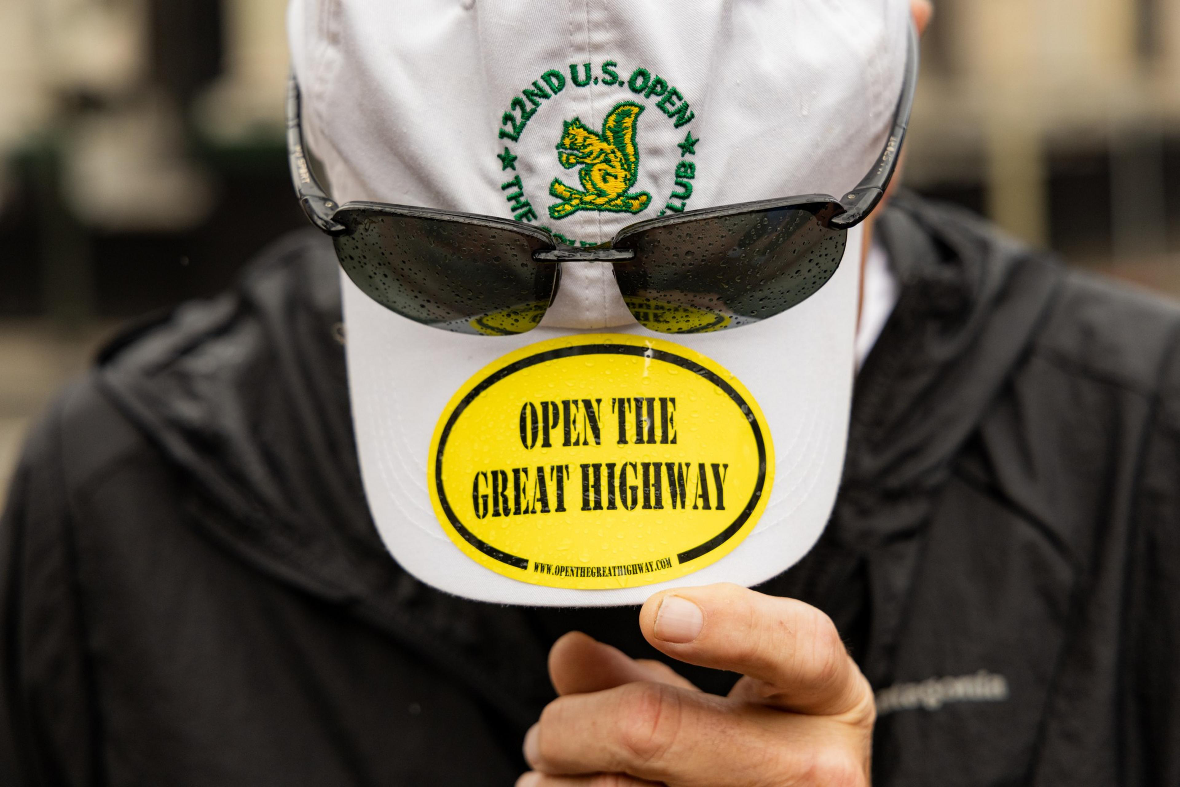 A person in a black jacket holds the brim of a white cap with dark sunglasses attached. The cap displays a yellow sticker reading &quot;OPEN THE GREAT HIGHWAY.&quot;