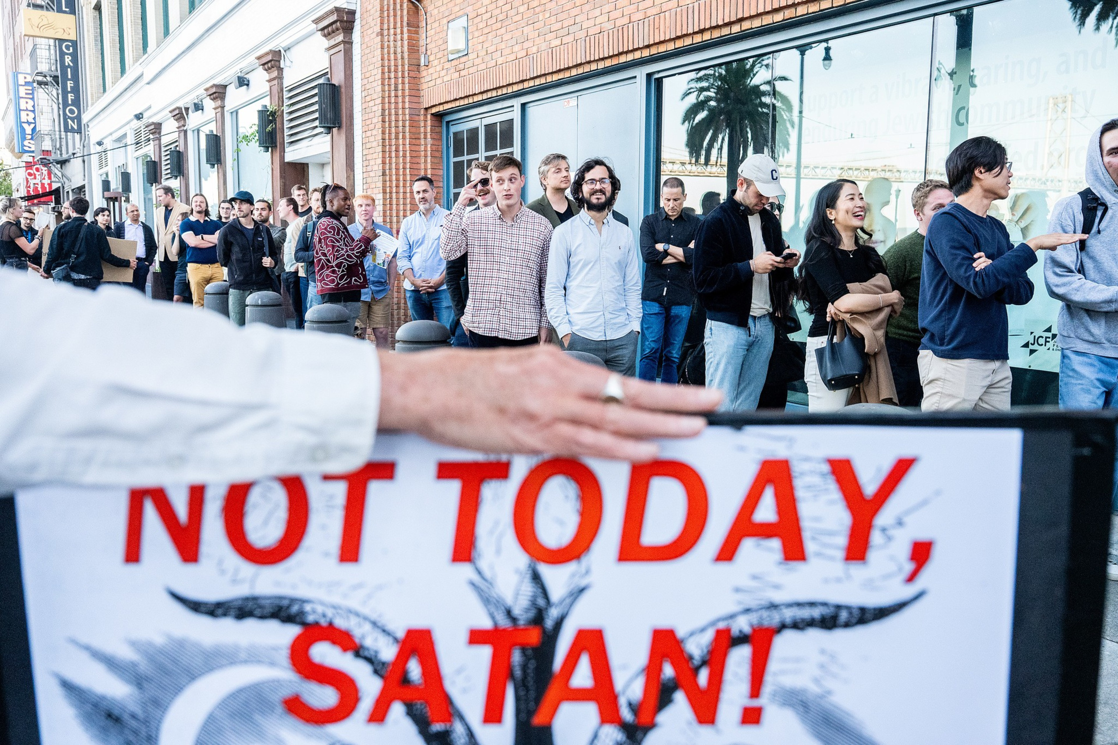 A diverse group of people wait in line outside a building while a hand holds a sign reading “NOT TODAY, SATAN!” in bold red letters.