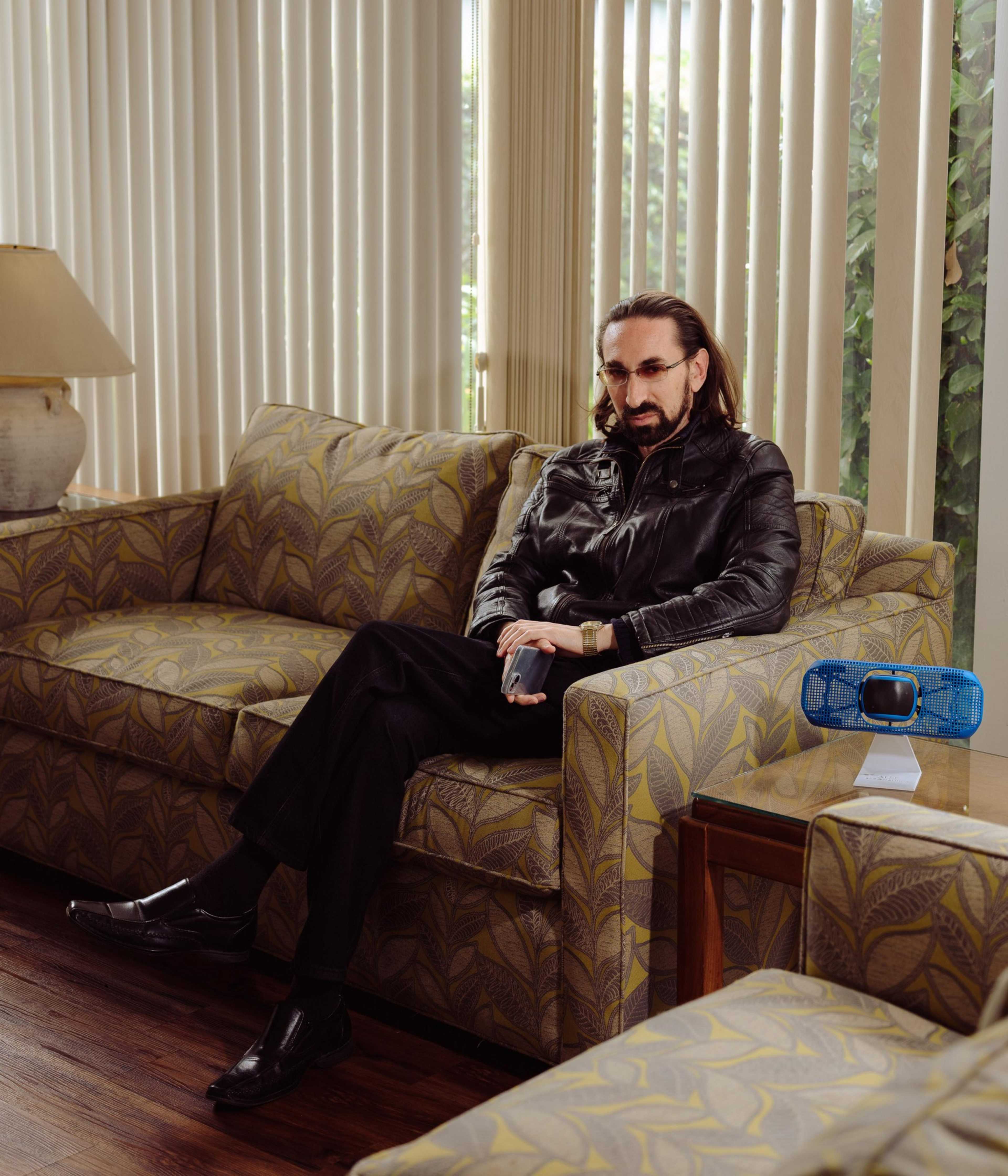 A person in a black leather jacket sits on a patterned sofa, holding a phone. There's a blue speaker on a side table, and blinds cover the windows.