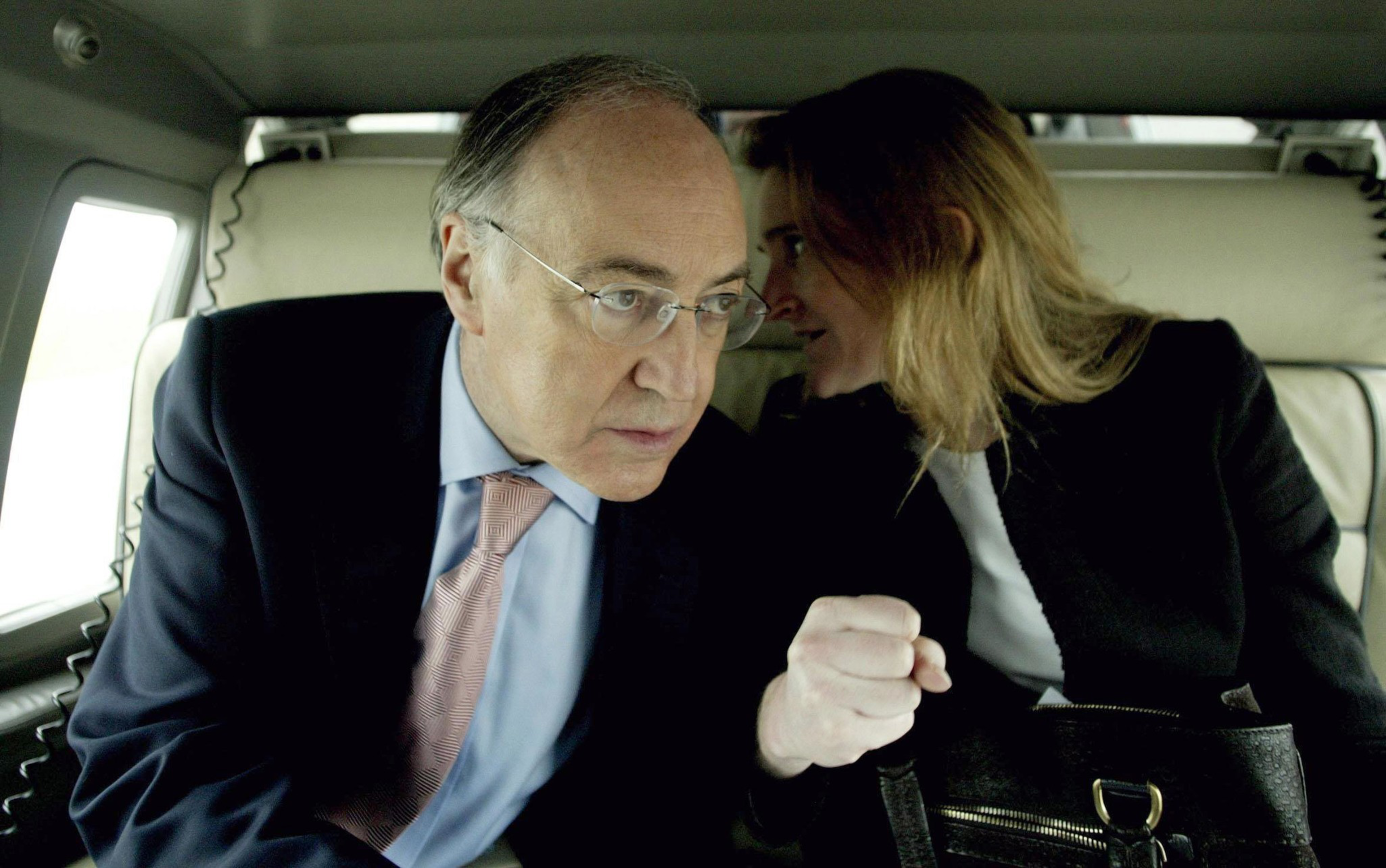 A woman in a black jacket whispers into the ear of a serious man in glasses and a suit inside a car.
