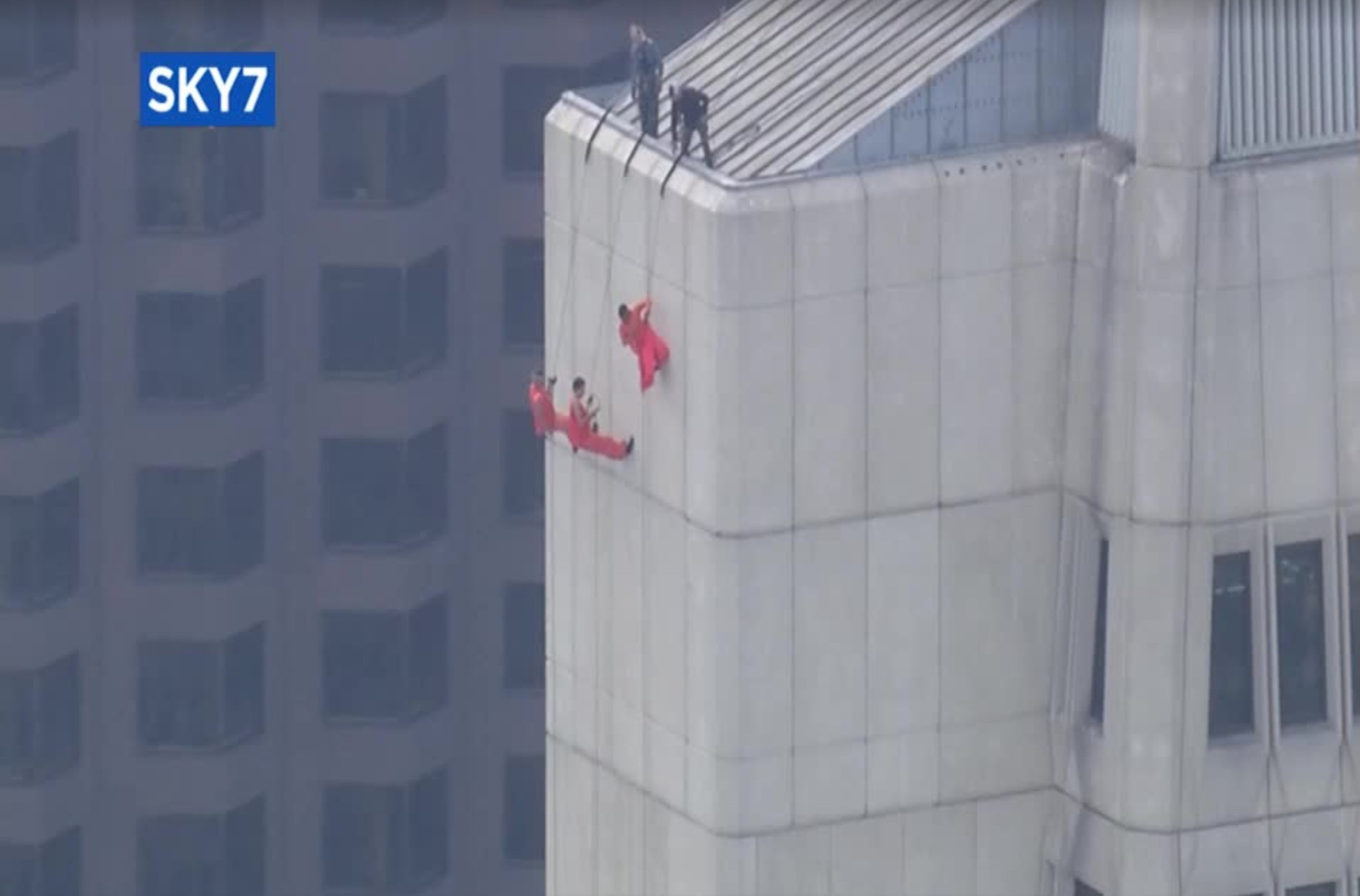 Three people wearing loose orange fabric sway and cling to cables held and maintained by three other dark-clothed figures atop a tall building's roof.