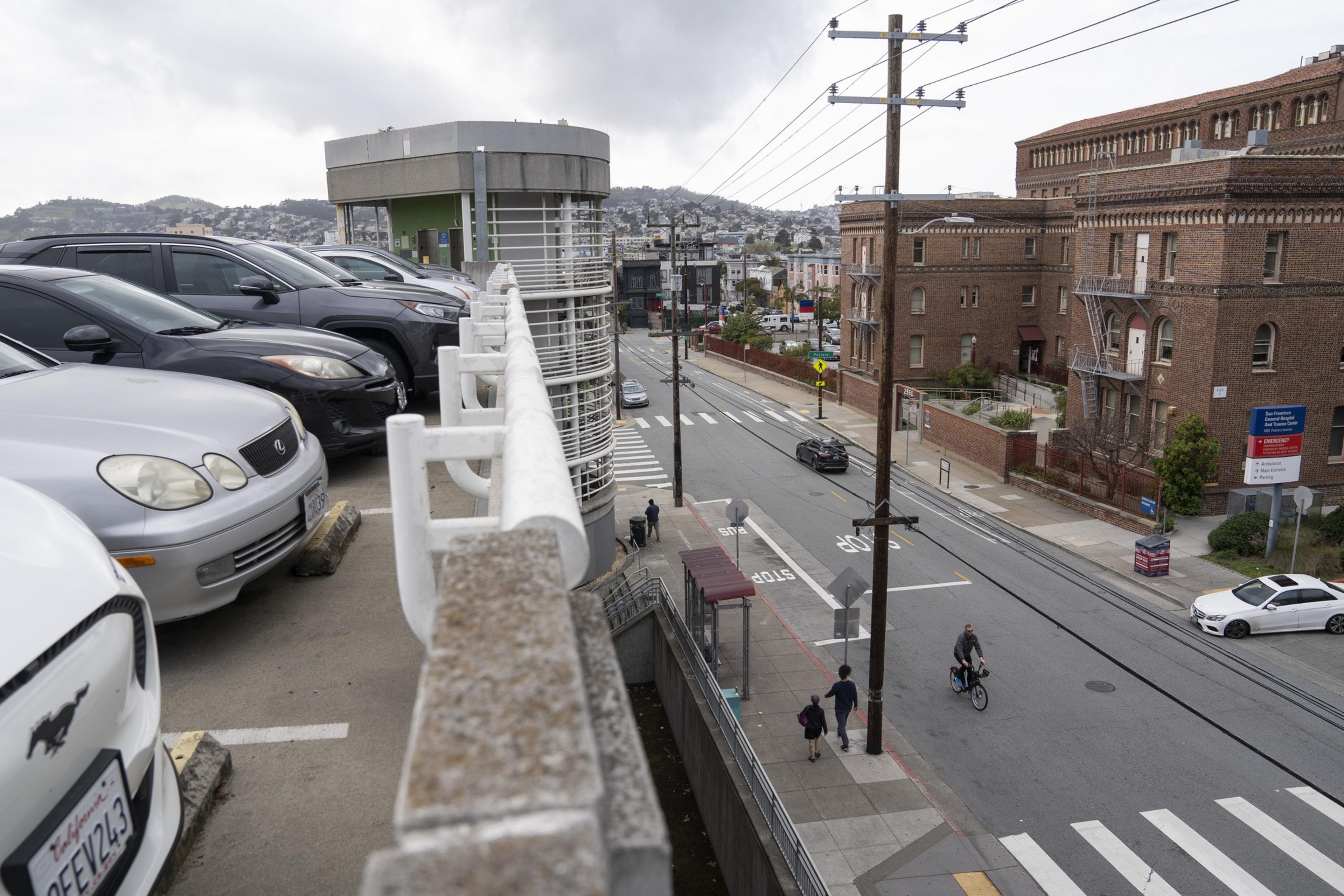 The image shows cars parked on a ramp, with a street below featuring cyclists and pedestrians. There's a red brick building and a backdrop of hills and cloudy skies.