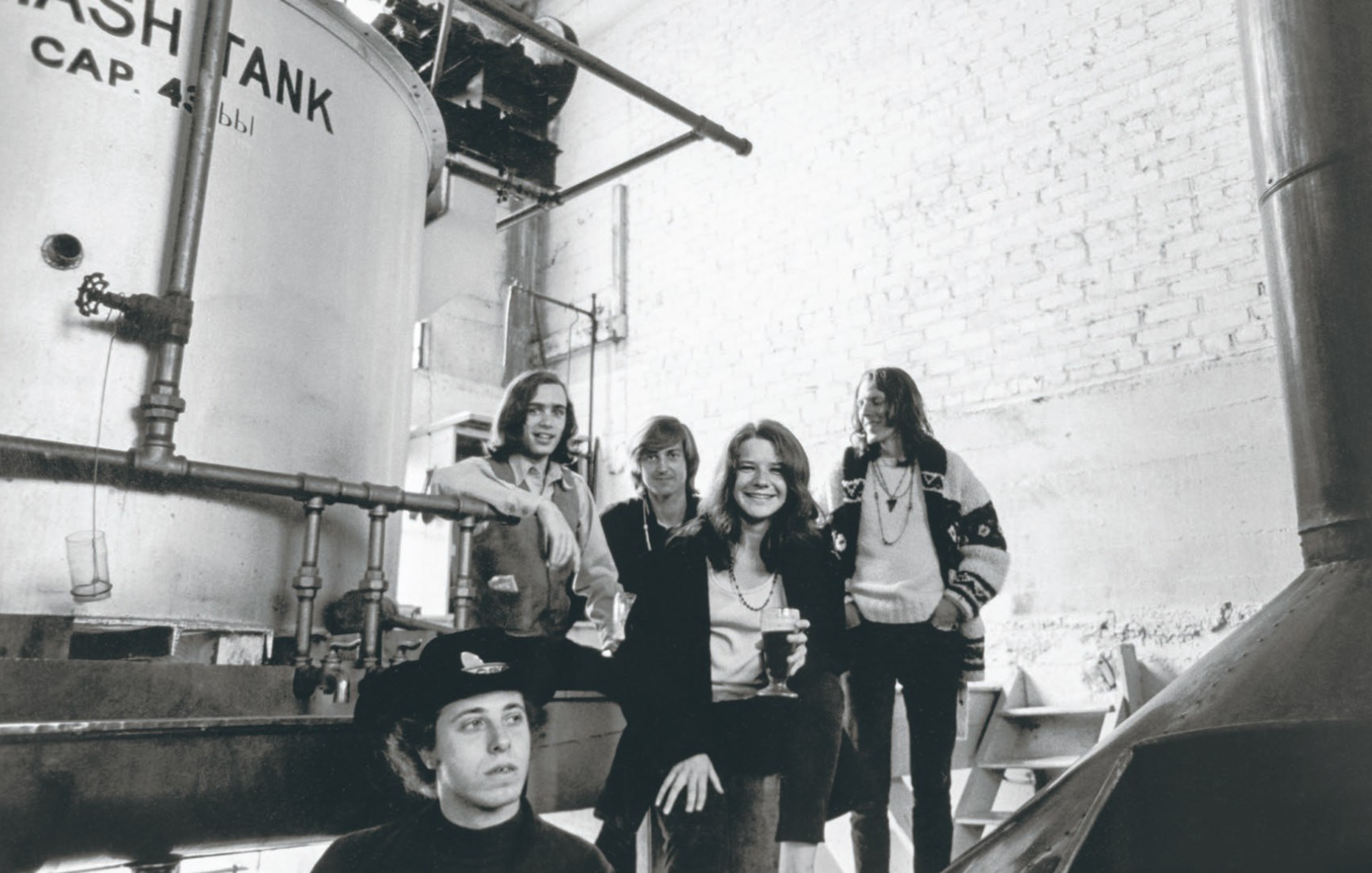 Five people pose casually in a brewery with large metal tanks, one person seated in front wearing a hat and others standing behind smiling and holding a drink.