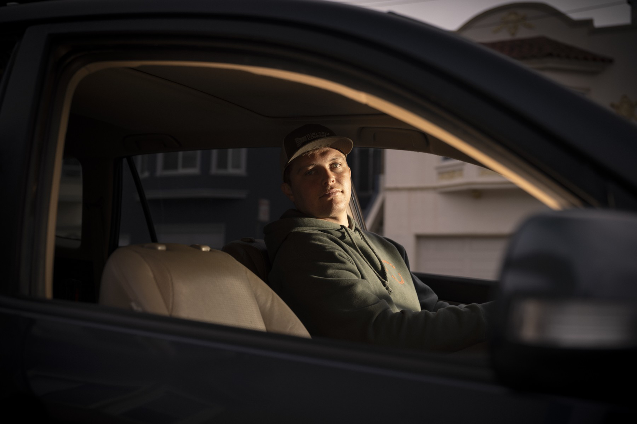 A person in a cap and hoodie sits in the driver's seat of a vehicle, looking out. The lighting is soft, highlighting their face, with buildings visible outside.