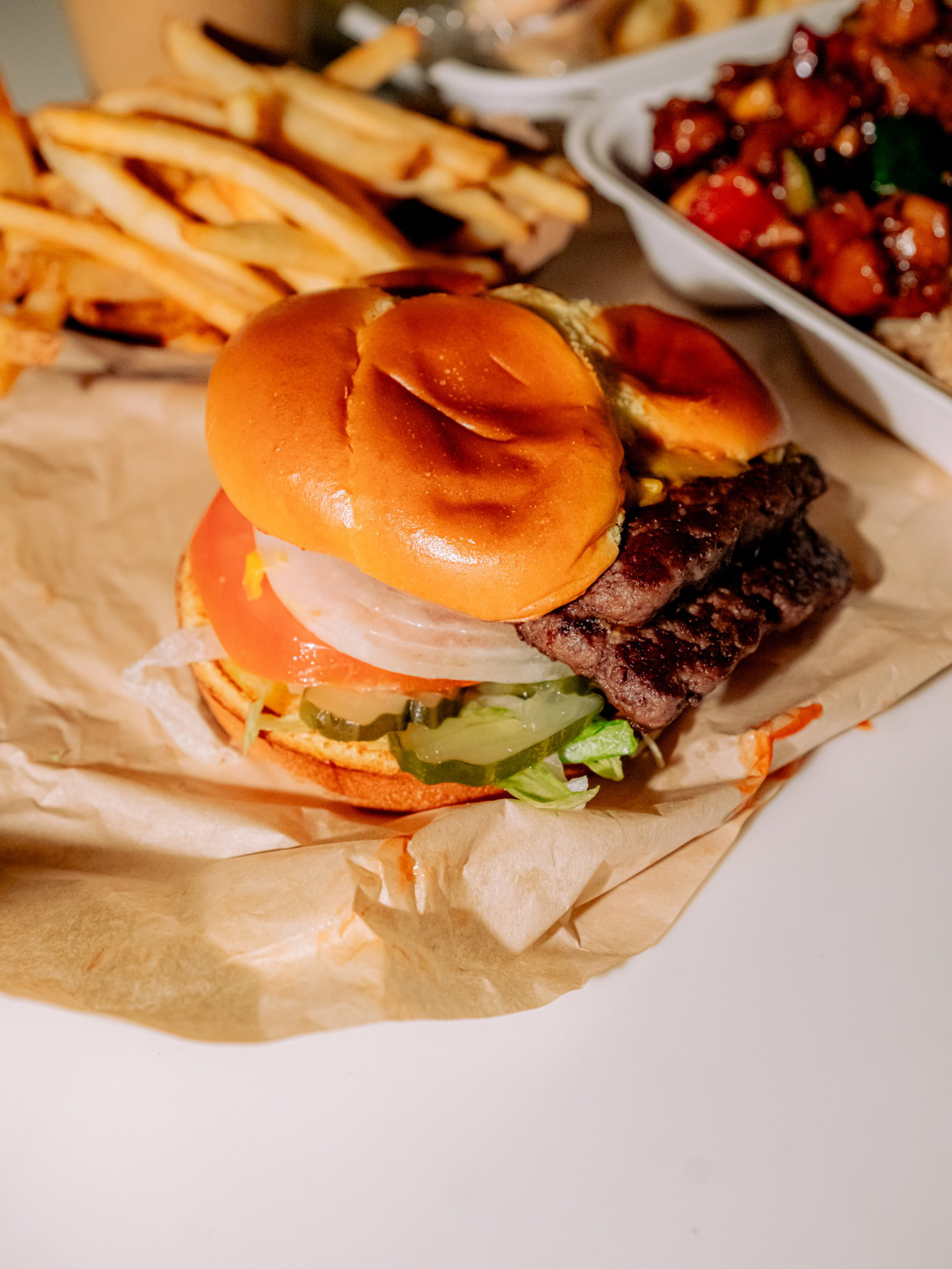 A cheeseburger with lettuce, tomato, onions, and pickles on a toasted bun, served with a side of French fries and a container of stir-fried food.