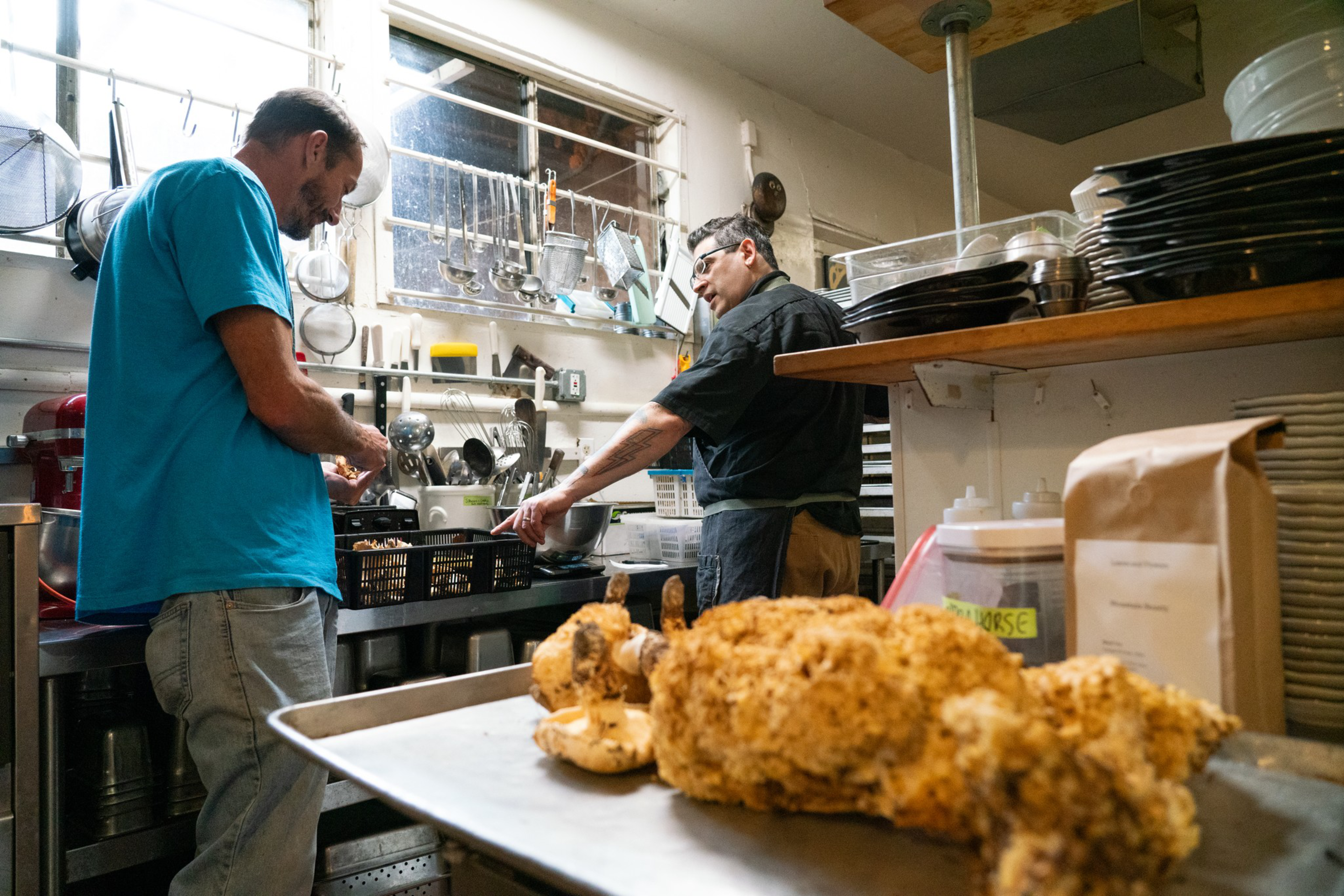 A man speaks to another man in a kitchen setting with wild mushrooms in the foregorund.