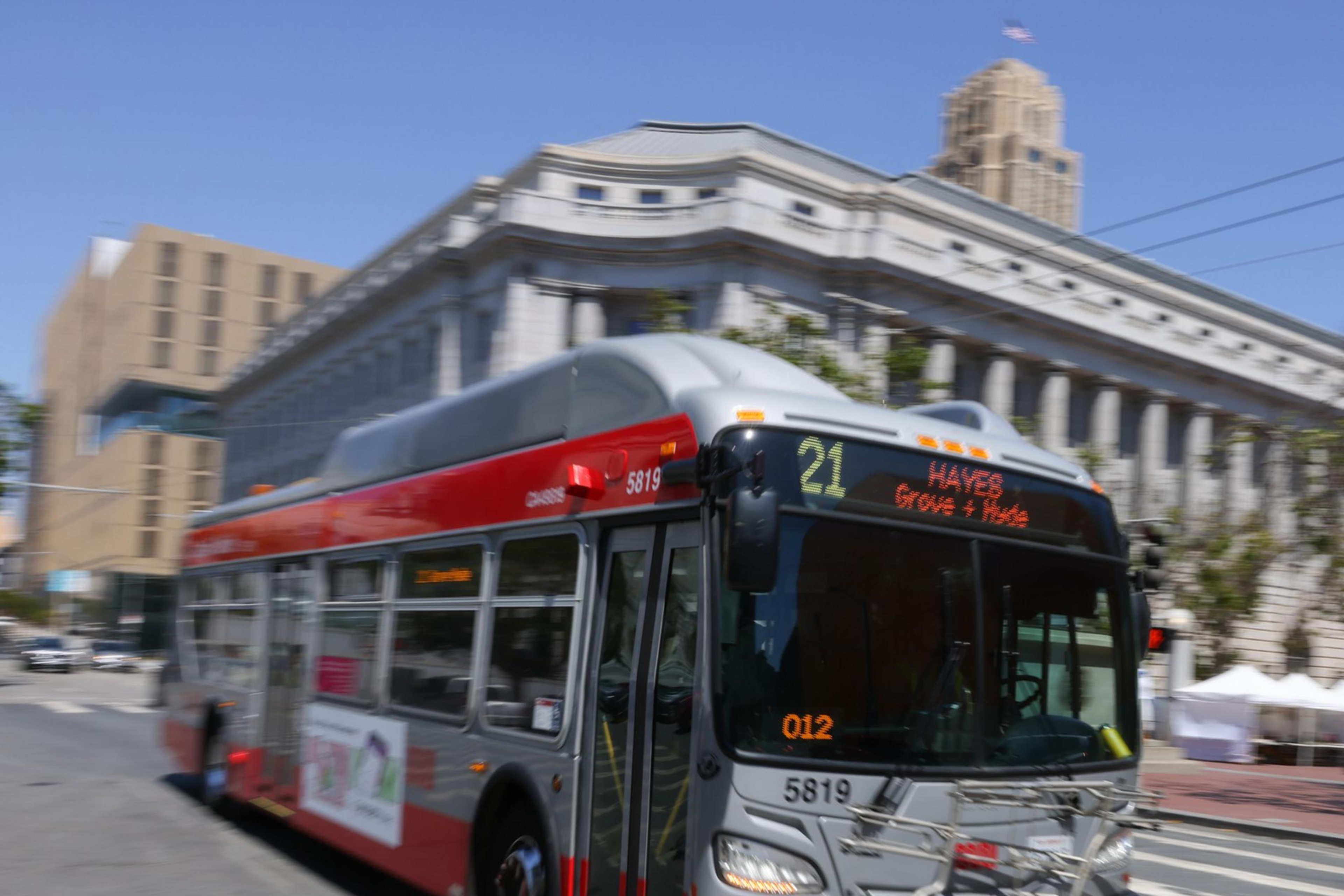 A gray and red city bus, marked route 21 to Hayes, moves along a crosswalk. It passes a large, classical-style building on a sunny day.