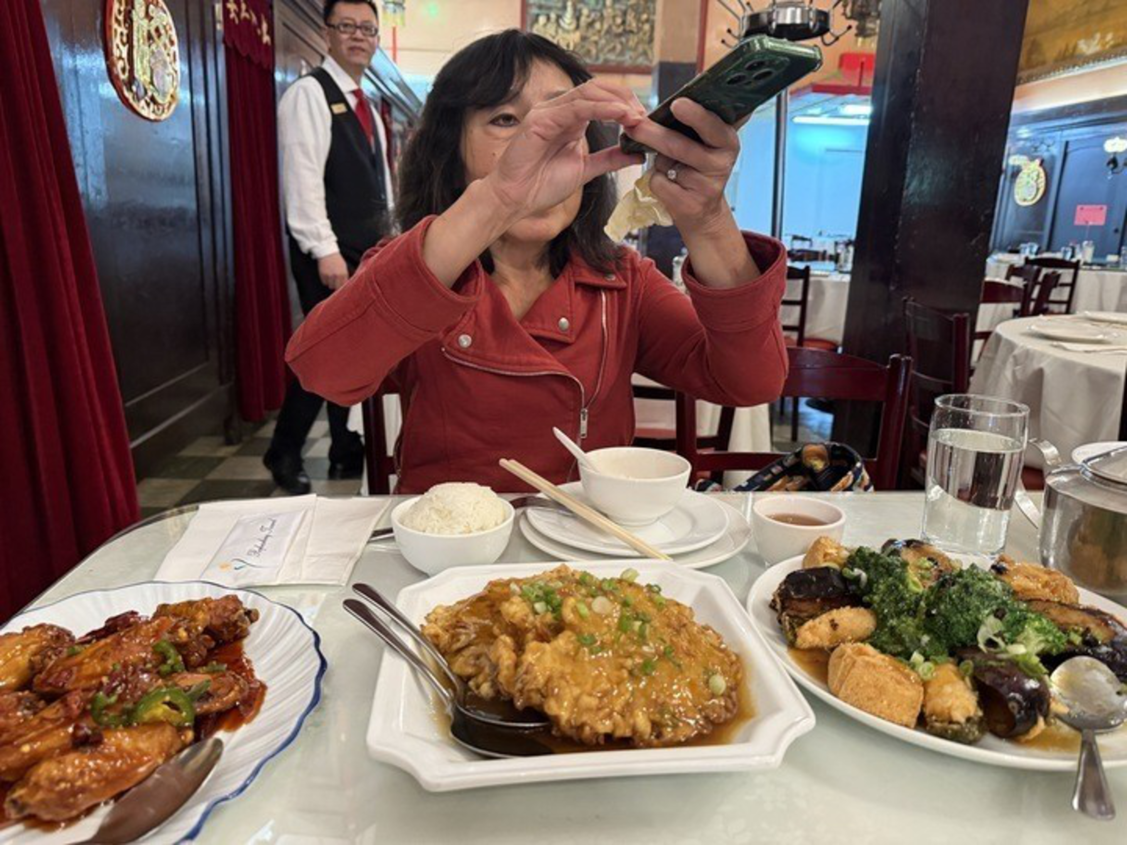 A woman in a red jacket takes a photo of a table with plates of Chinese food, including shrimp, an egg dish, and vegetables with tofu.
