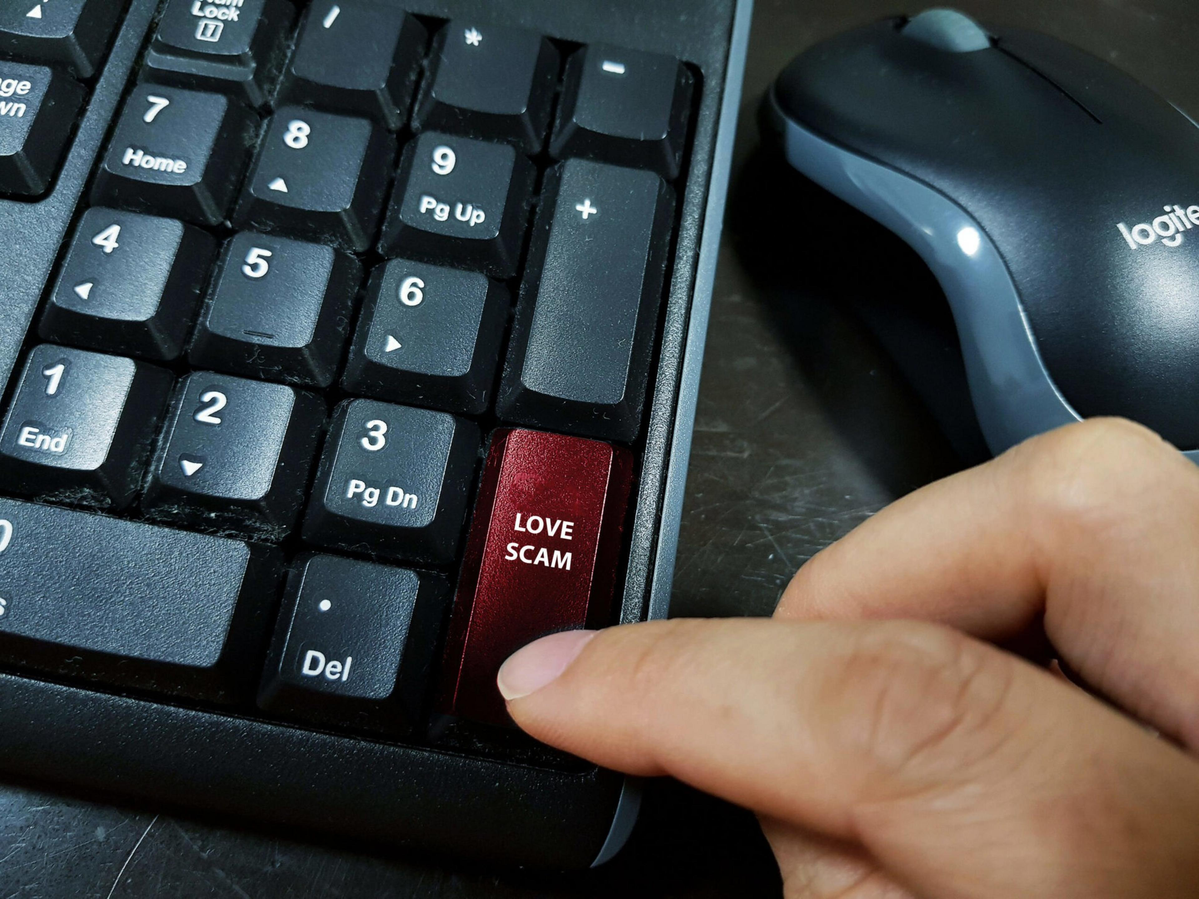 A red key labeled "LOVE SCAM" replaces the Enter key on a black numeric keypad, with a finger about to press it and a Logitech mouse nearby.