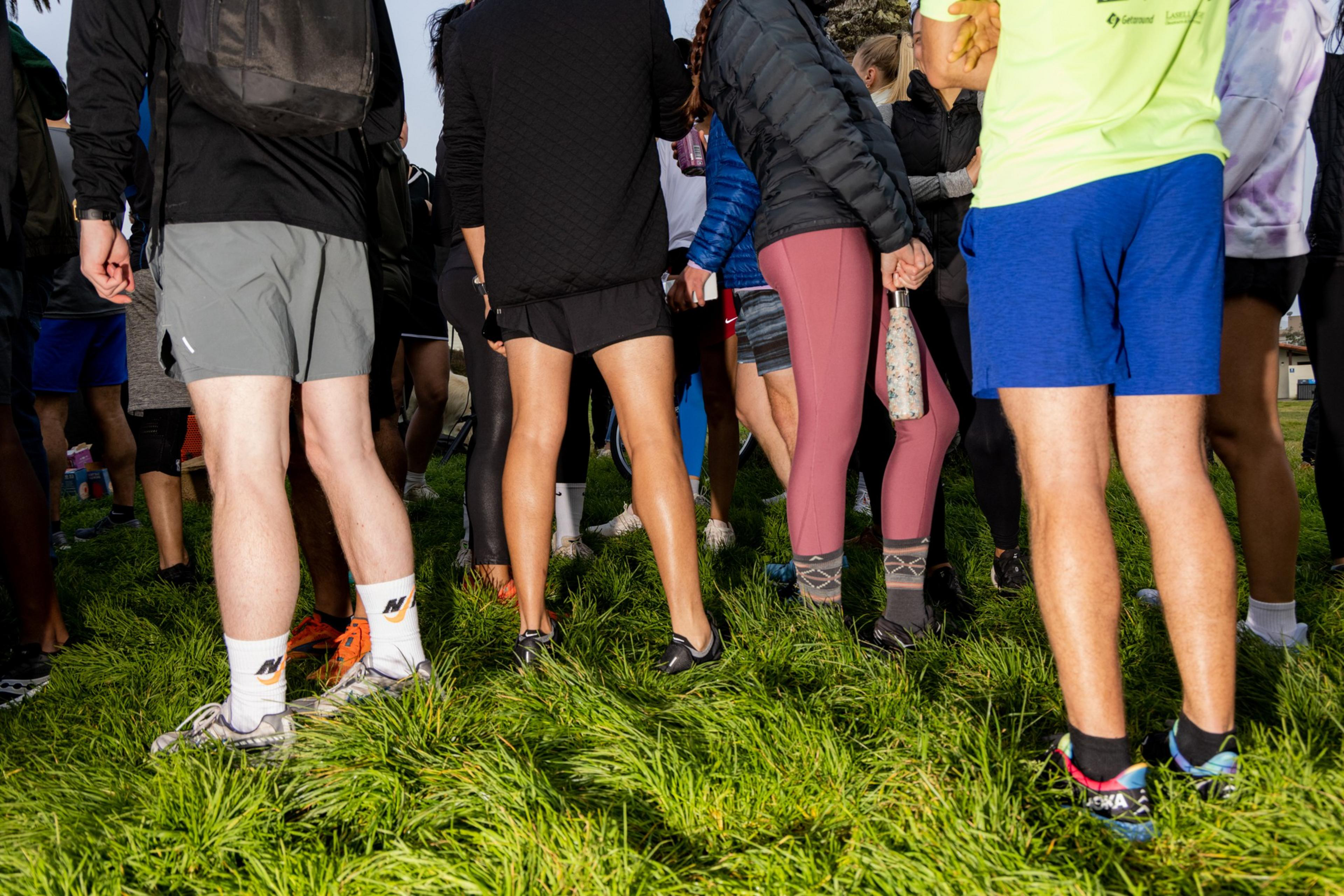 A group of people in athletic wear are standing on green grass. Their legs and lower bodies are mostly visible, showing a mix of shorts, tights, and sneakers.