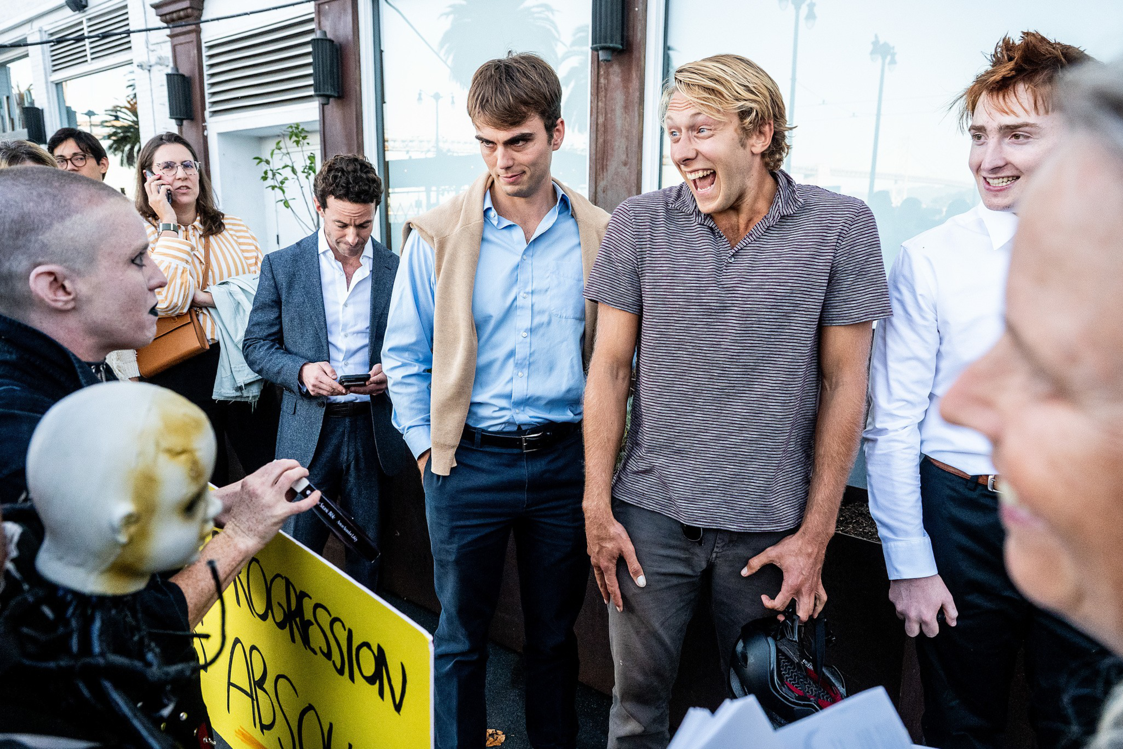 A group of young people stand outside, some smiling and one looking skeptical, while a person holds a yellow sign with partially visible text.