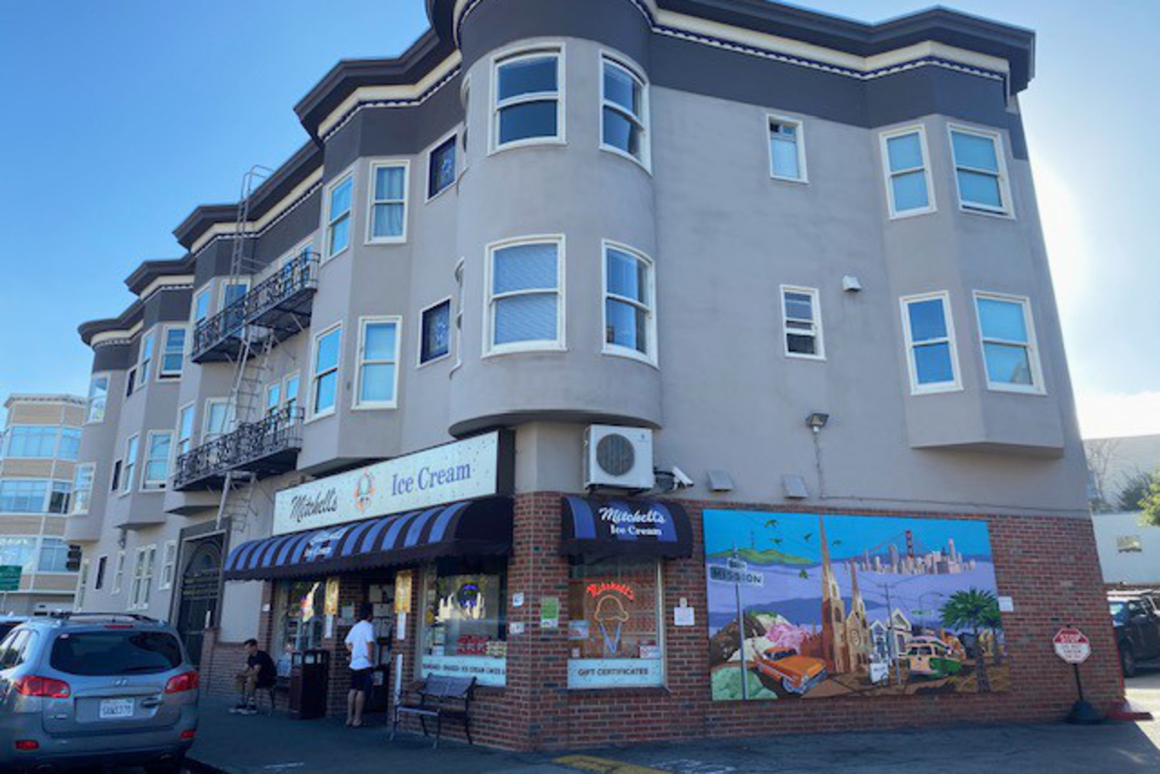 The image shows a beige, multi-story building with an ice cream shop on the ground floor. The shop has a sign and a striped awning, with a mural painted on the side wall.